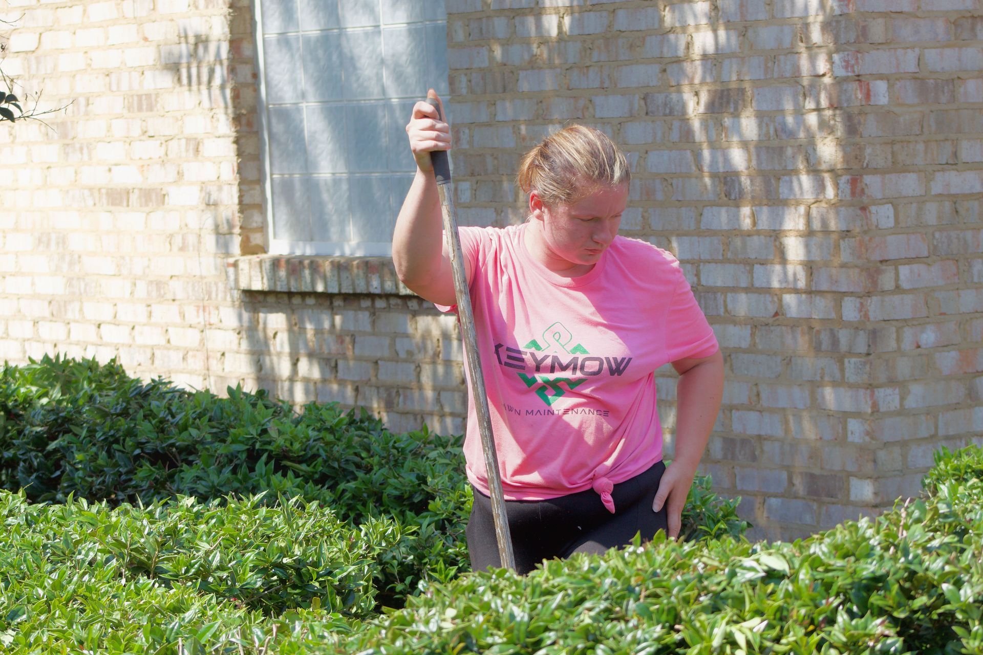 Person in pink shirt holding a long tool while standing in garden beside a brick wall