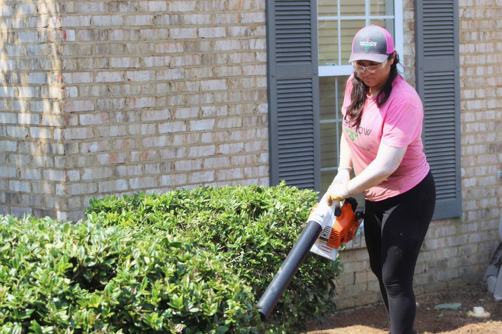 Person using a leaf blower near a brick building with green shrubs and shuttered windows