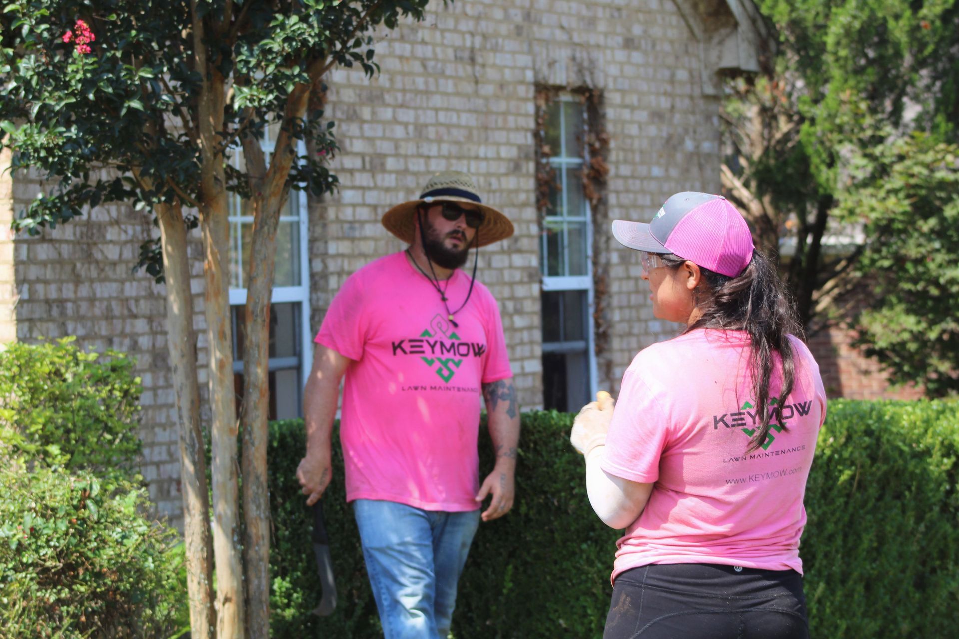 Two people in pink shirts talking outdoors near a brick building and trees.