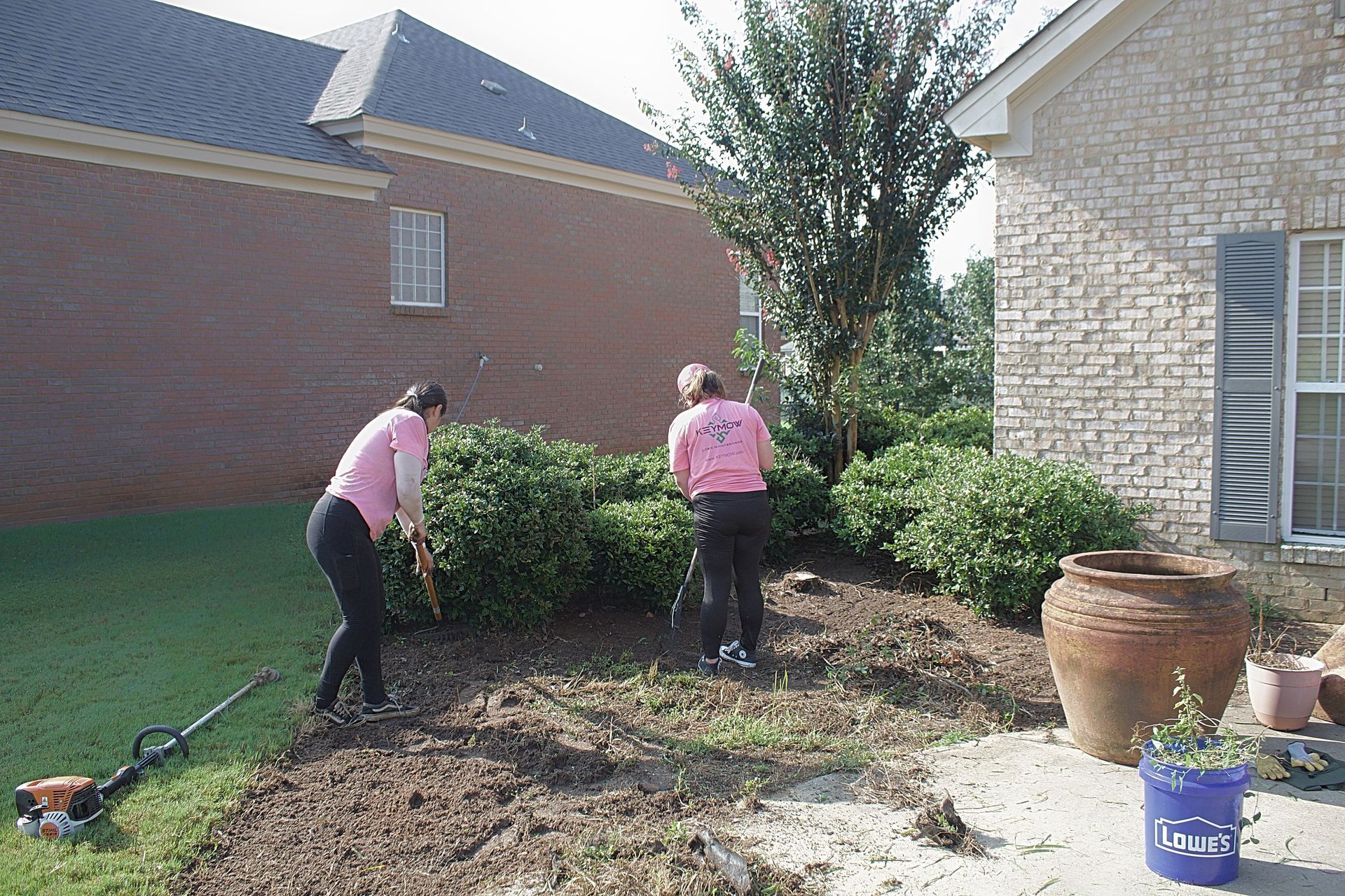 Two landscapers in pink shirts trimming a hedge beside a brick house and stone patio