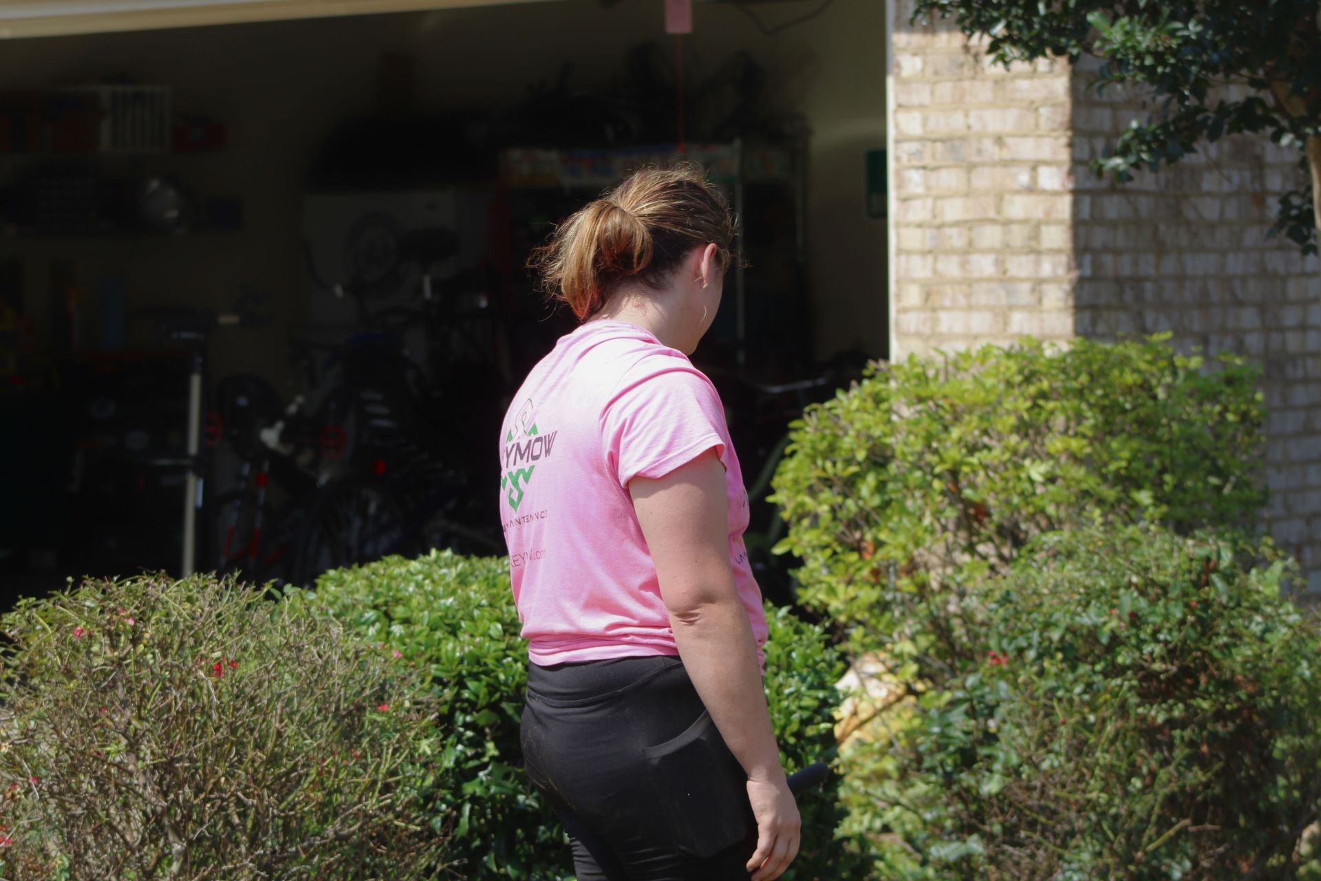 Person in a pink shirt standing near bushes outside a garage doorway