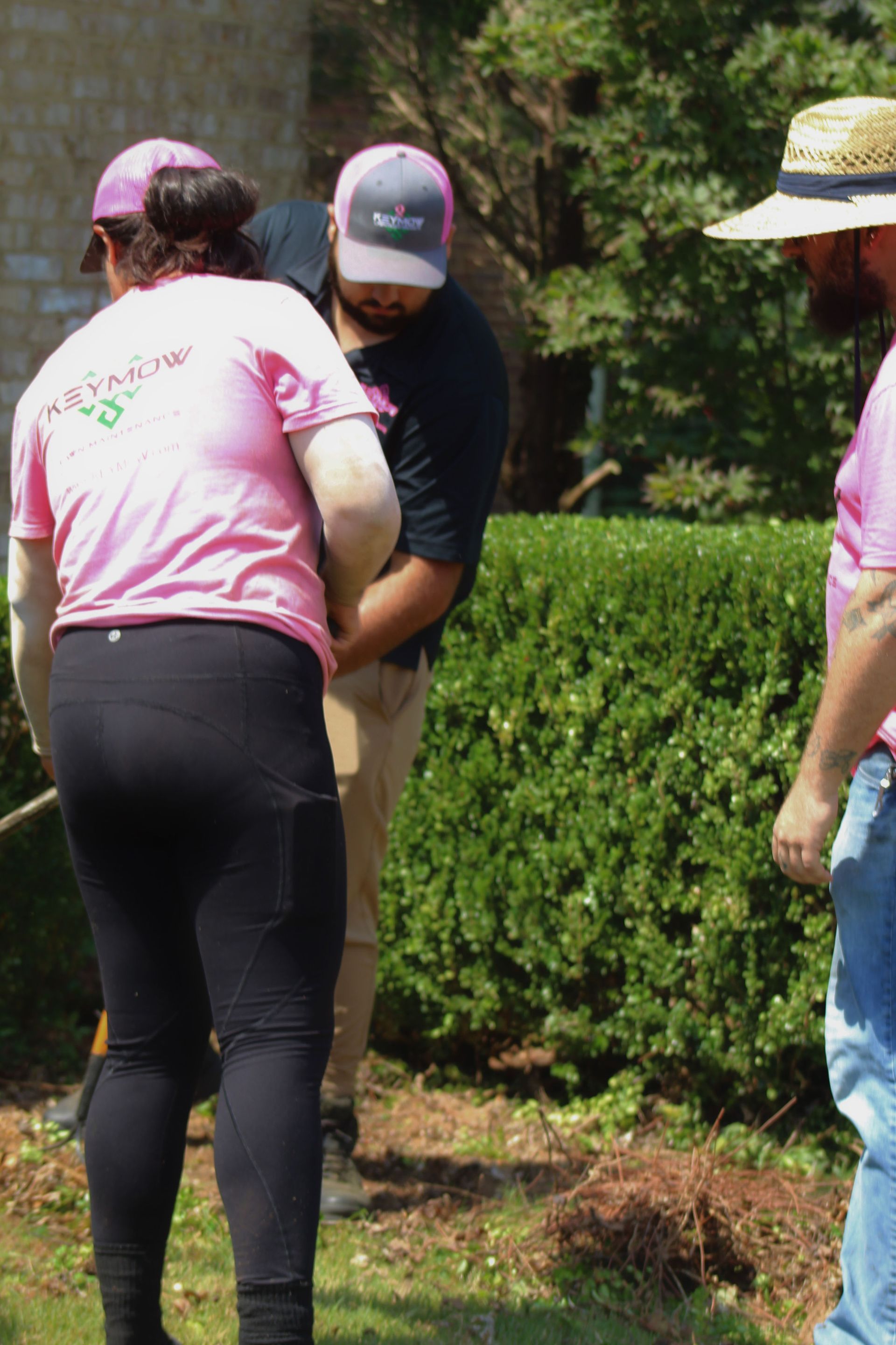 Three people in pink shirts and caps standing outdoors near a hedge and trees
