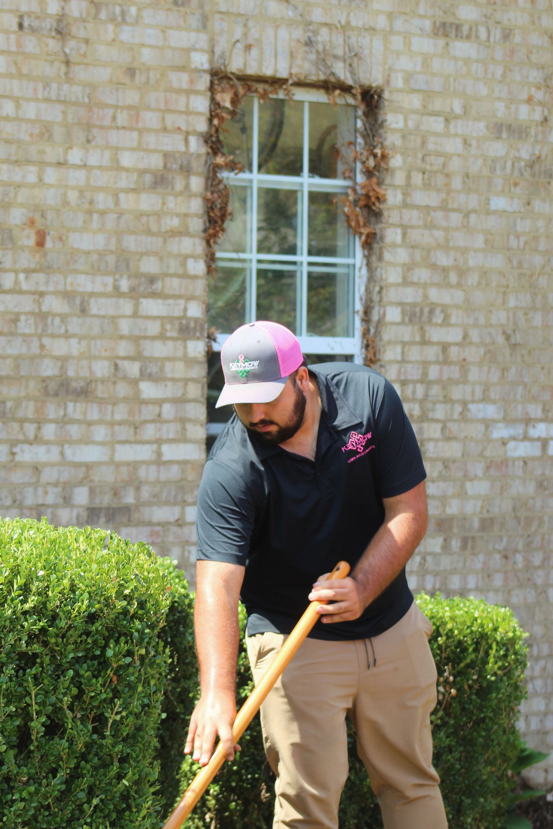 Man trimming a hedge with a long-handled tool in front of a brick building