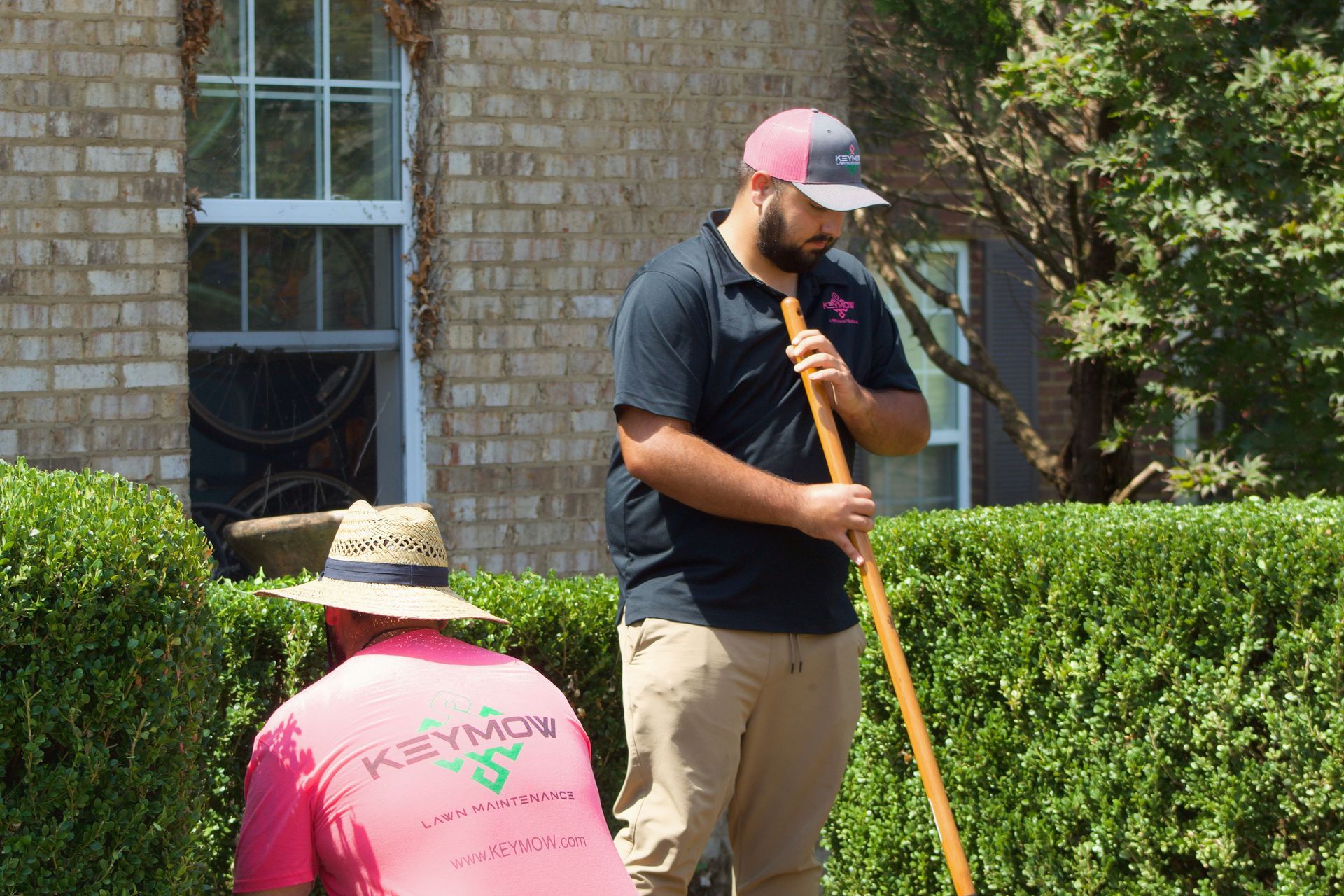Two workers gardening outside a brick building, one standing with a tool and one kneeling by a hedge.
