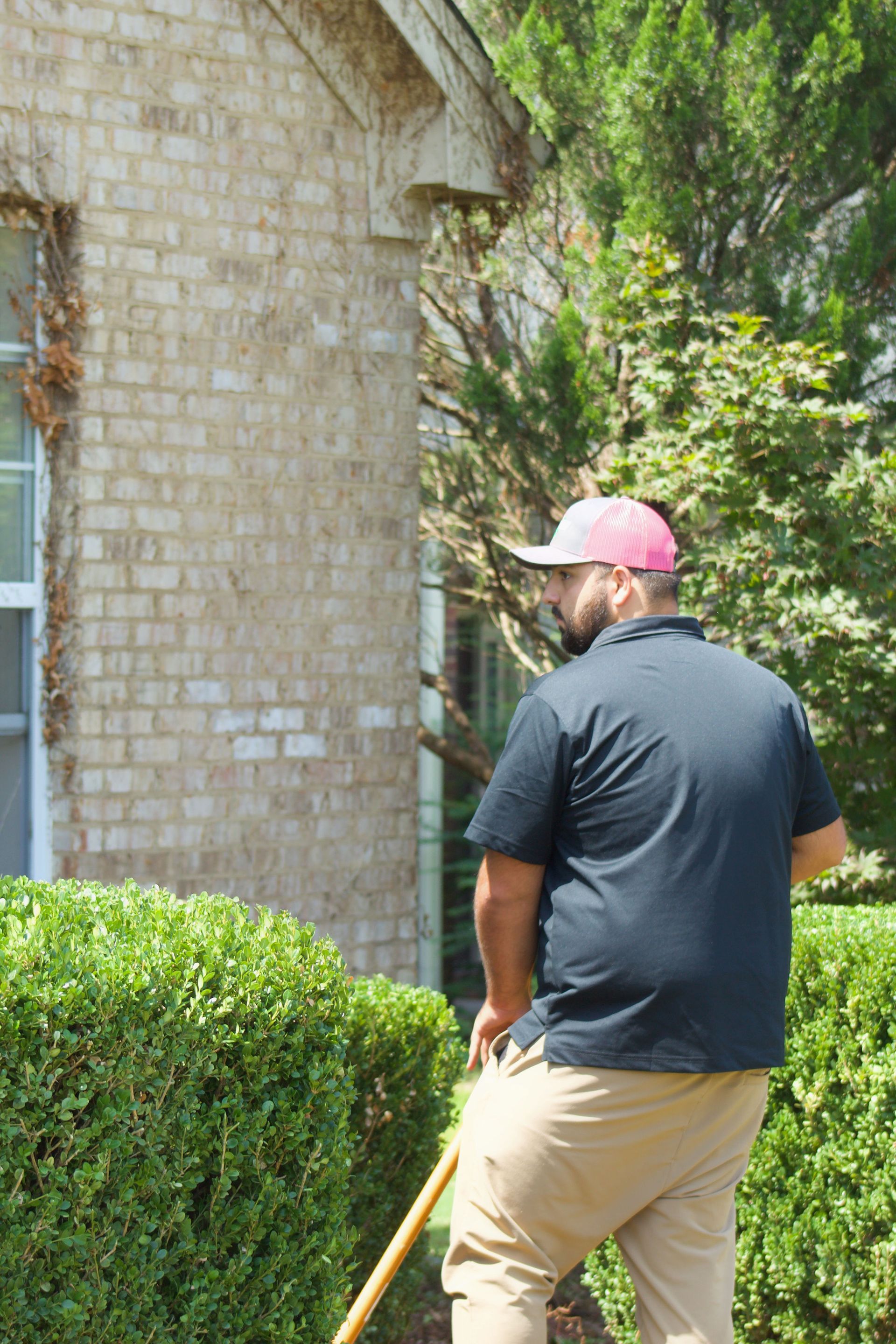 Person in pink cap and dark shirt using a shovel beside a brick house and green hedge.