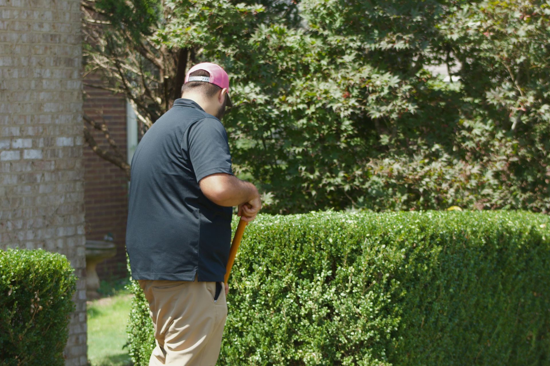 Person watering a hedge in a garden beside a brick house, holding a yellow hose.