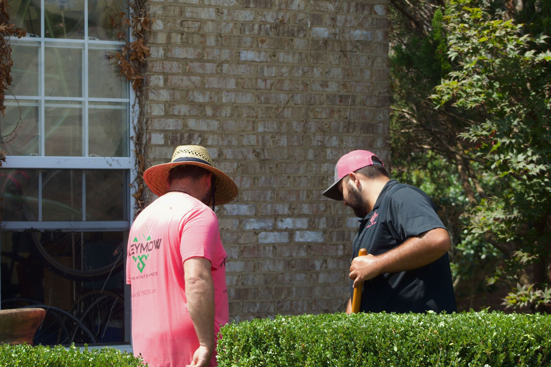Two people in sun hats stand by a hedge near a brick house, one digging with a tool.