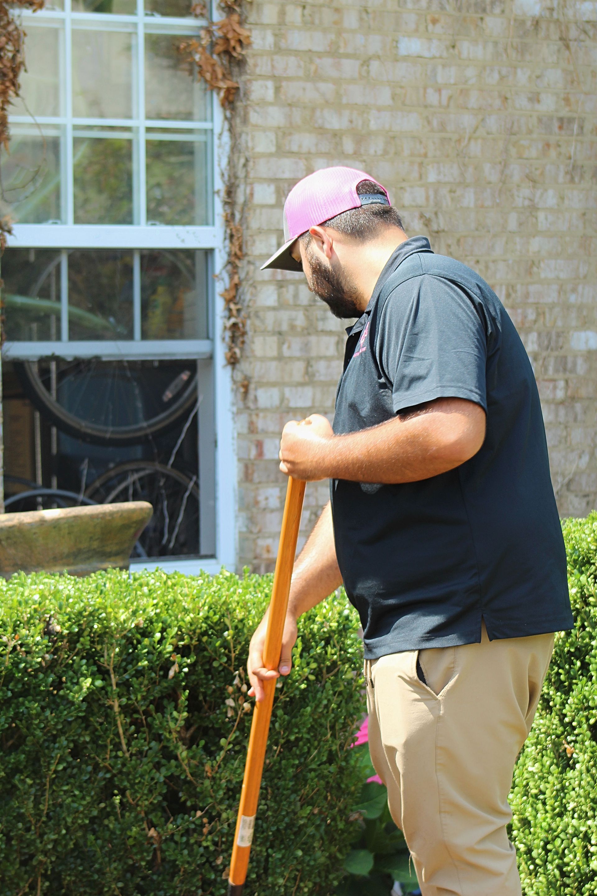 Person in a pink cap using an orange tool beside a hedge outside a brick building.