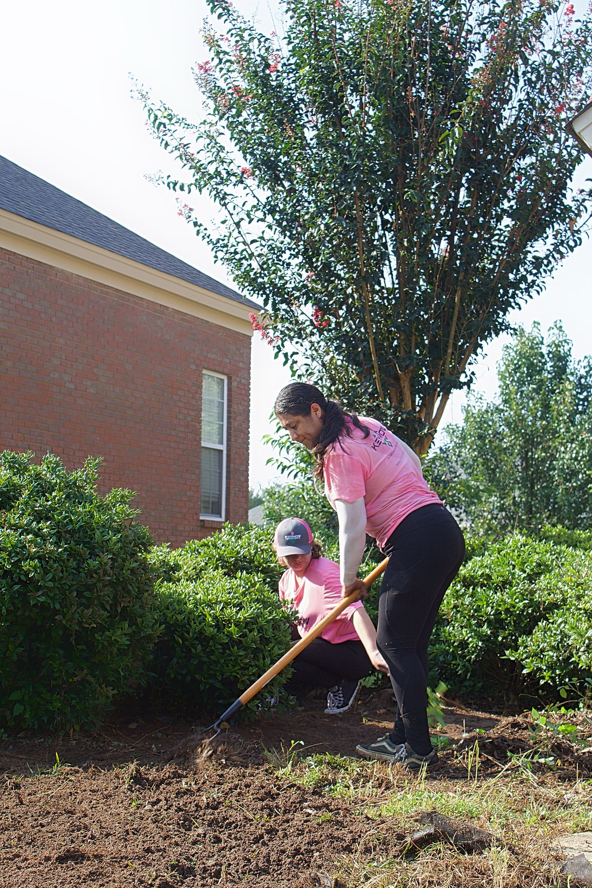 Two people gardening with a rake beside a brick building and trees