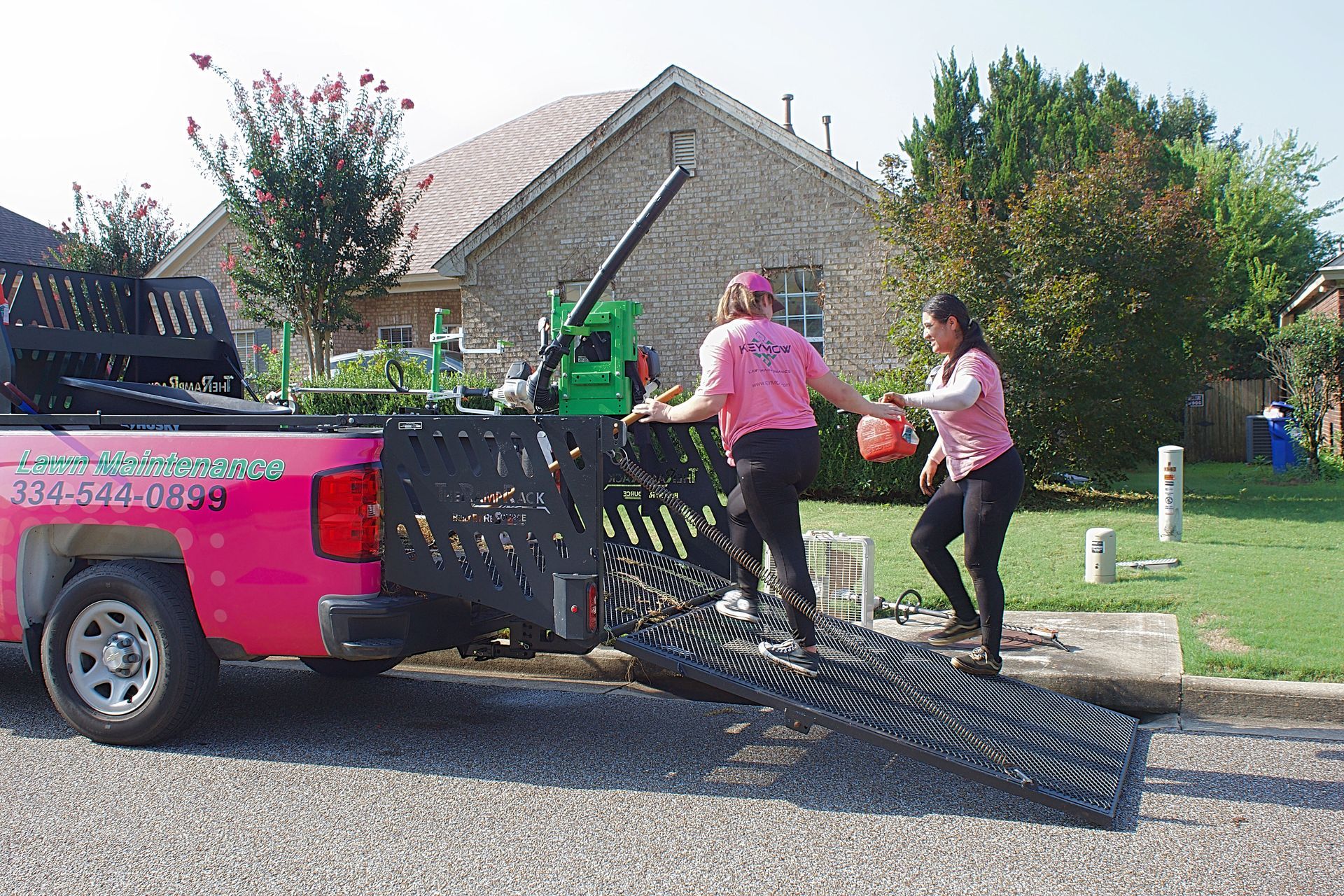 Two people loading a black metal ramp into a pink pickup truck in a residential driveway