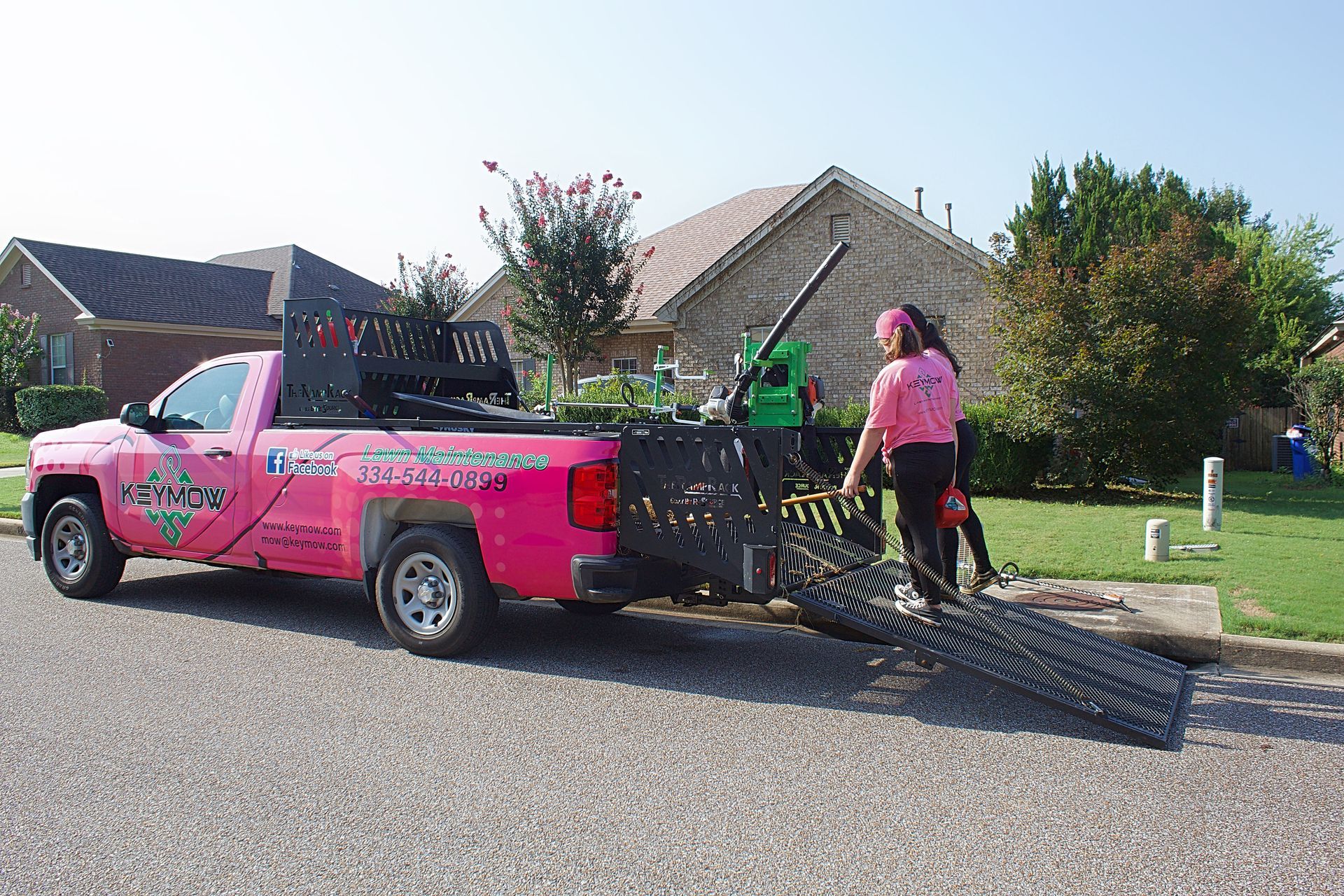 Pink pickup truck with a metal ramp, two workers loading equipment on a suburban street