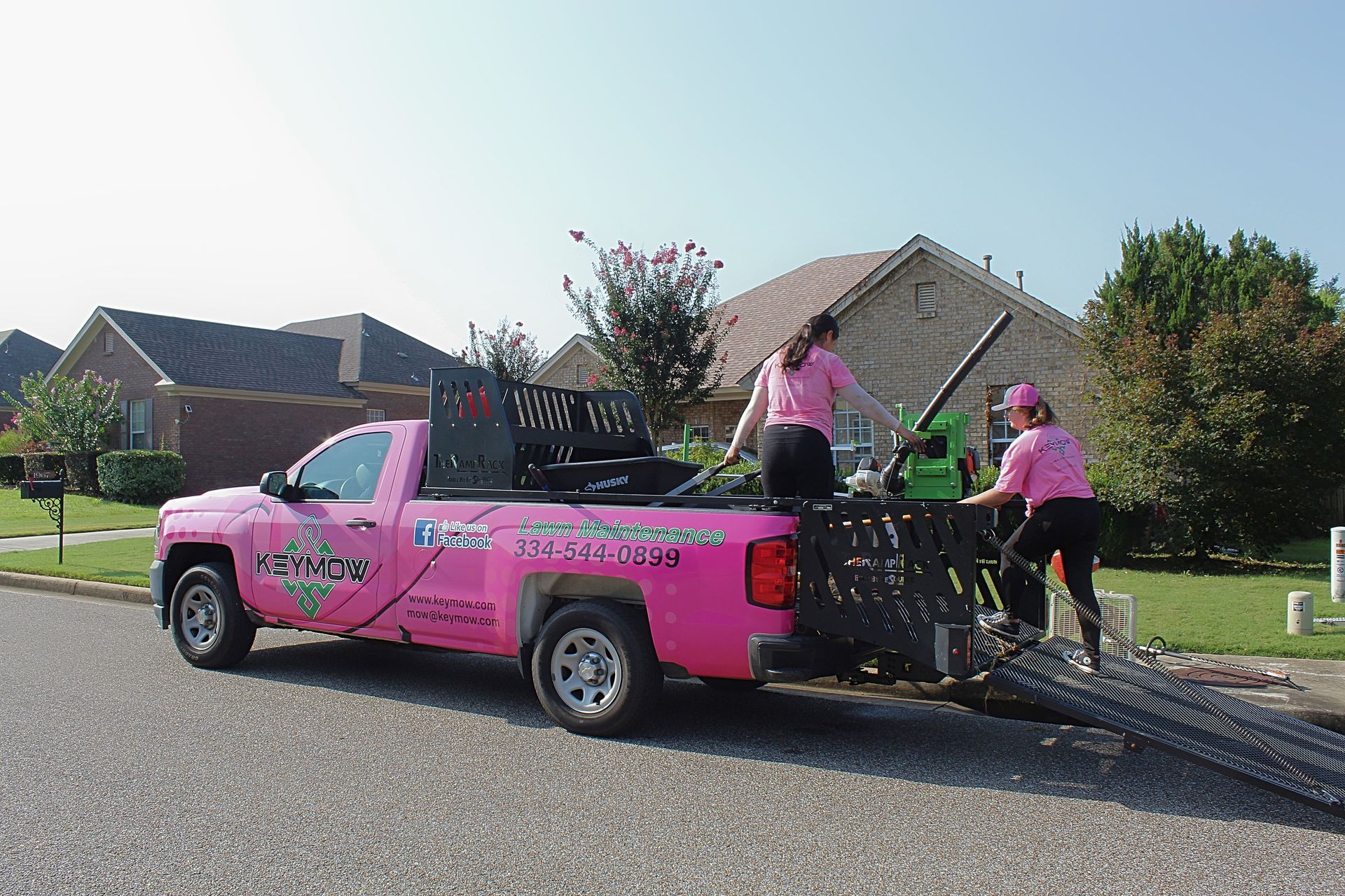 Pink pickup truck unloading long black ramps on a suburban street, with workers beside it.