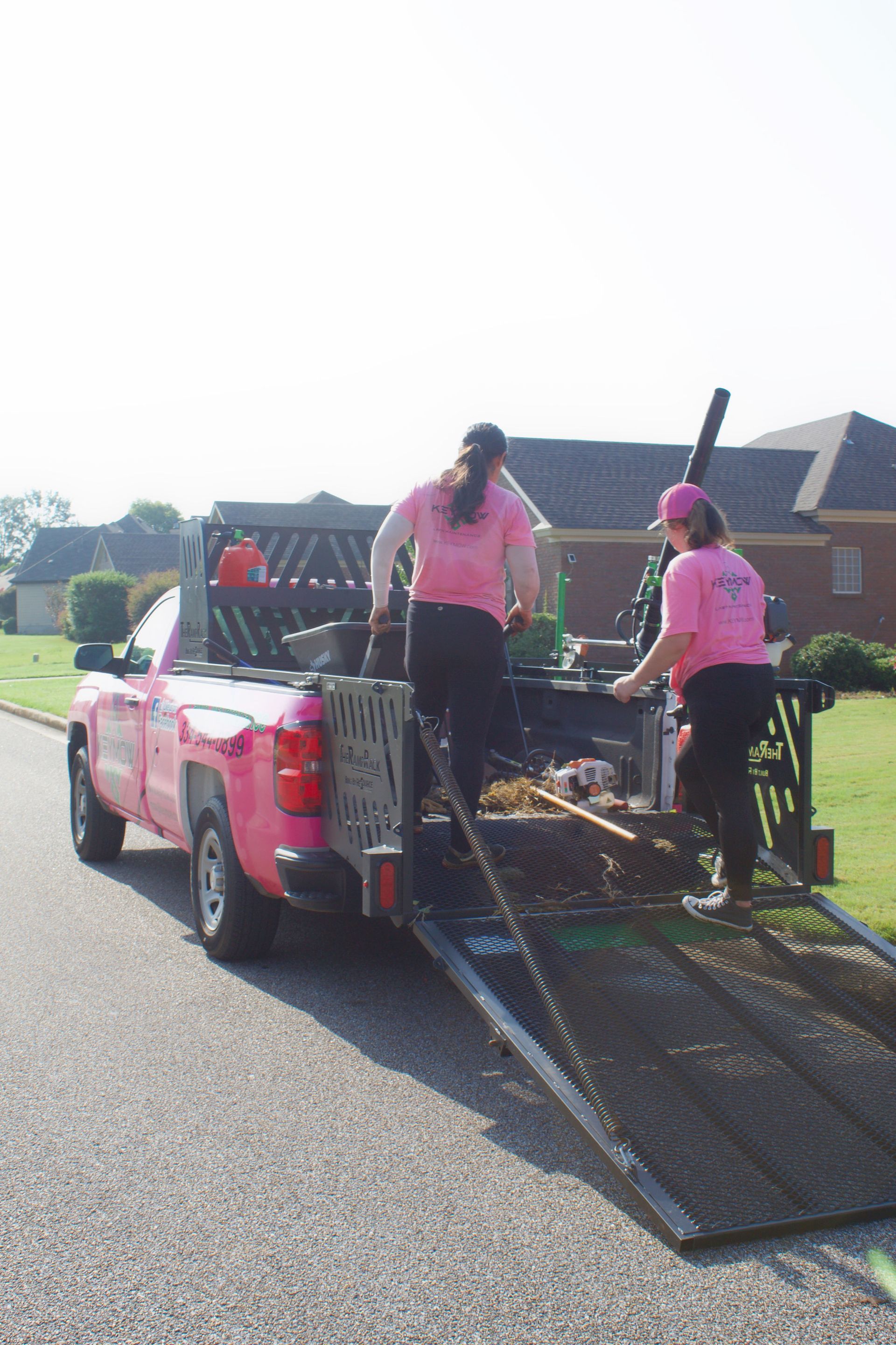 Two workers loading a red utility truck with a metal ramp on a residential street.