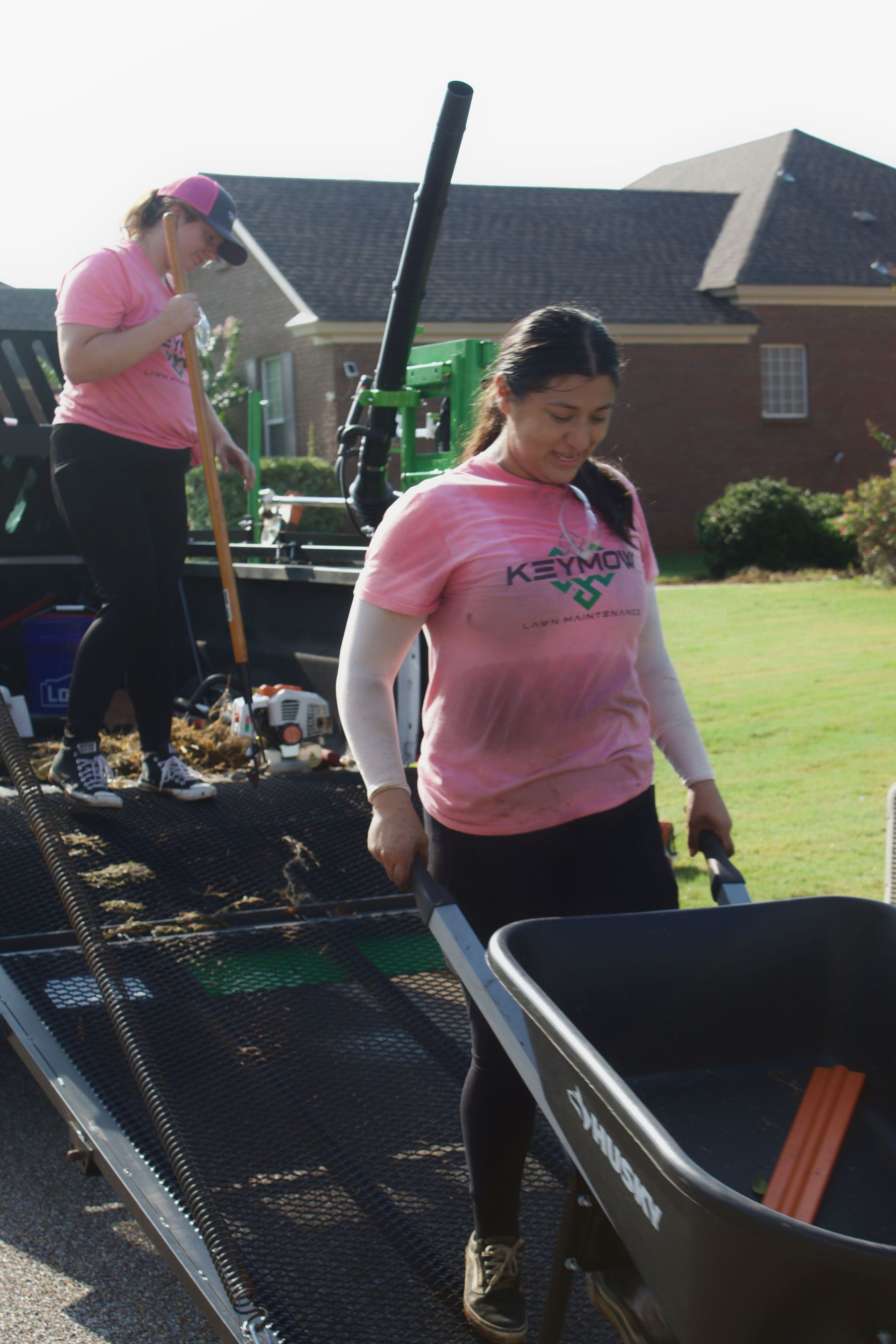 Two people in pink shirts moving wheelbarrows on a long ramp outdoors near a brick building.