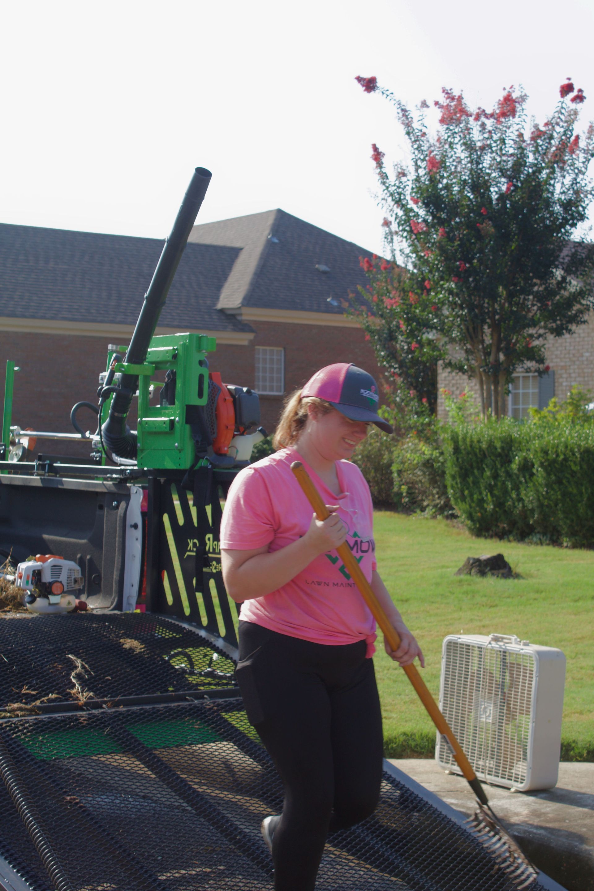 Person in pink shirt and cap walking beside a parked green utility truck in a suburban yard.