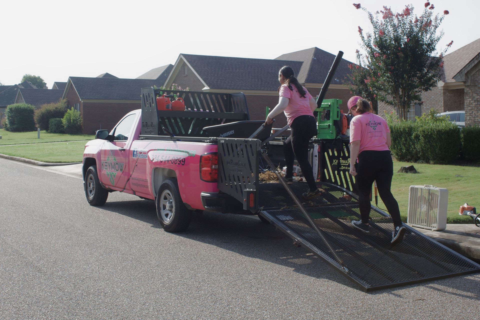 Pink truck with ramp in a driveway, with two people loading equipment into the back.
