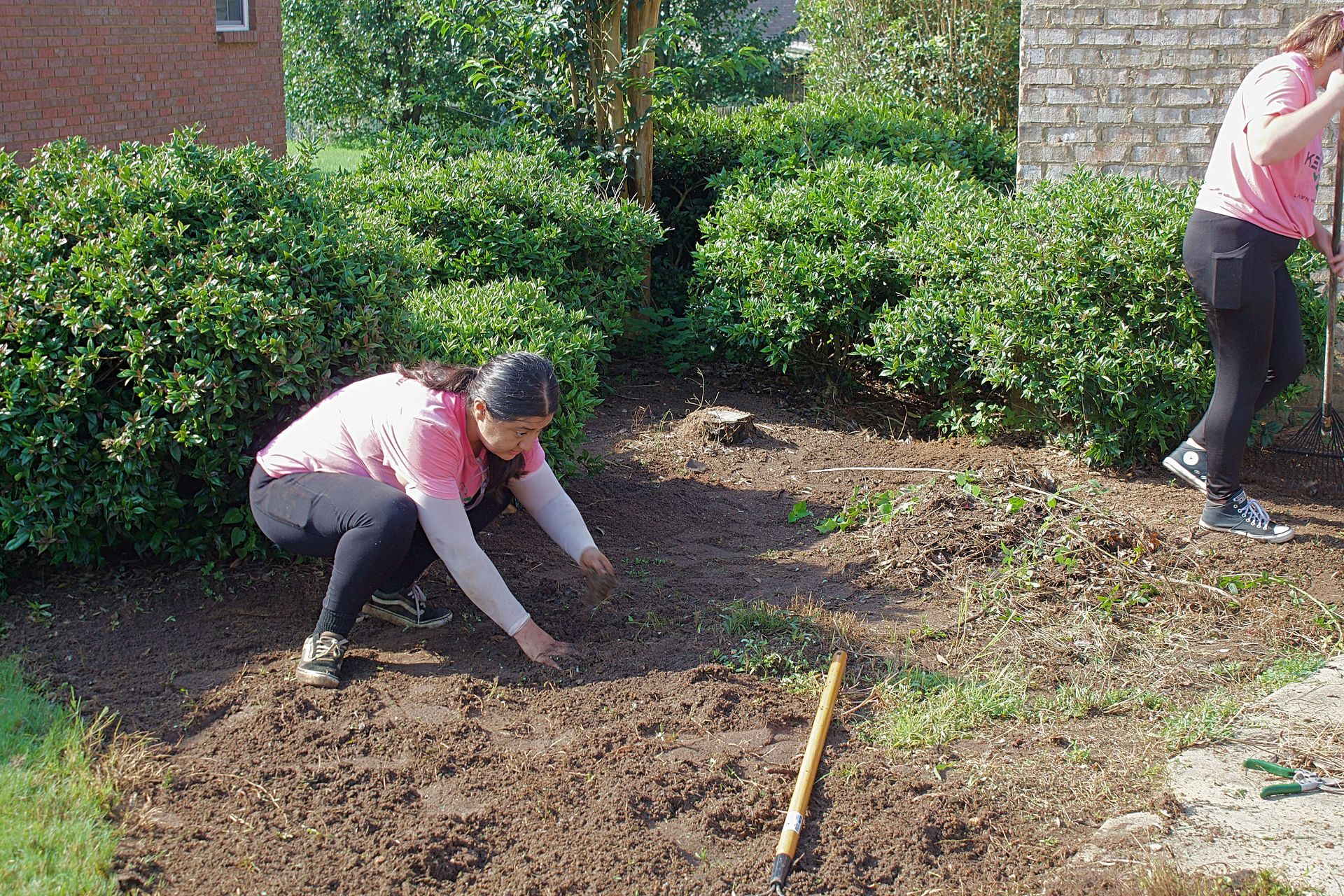 Two people planting or gardening in a mulched flower bed beside a brick house