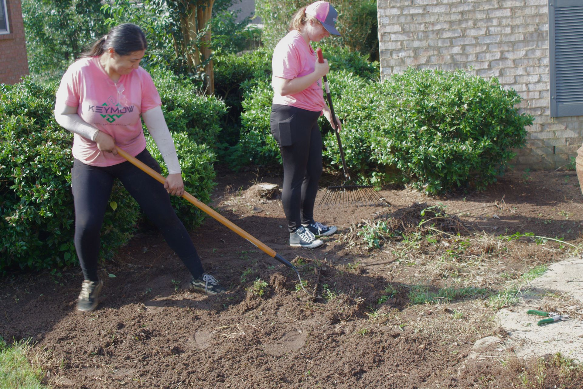 Two people in pink shirts raking soil in a garden bed beside a house.