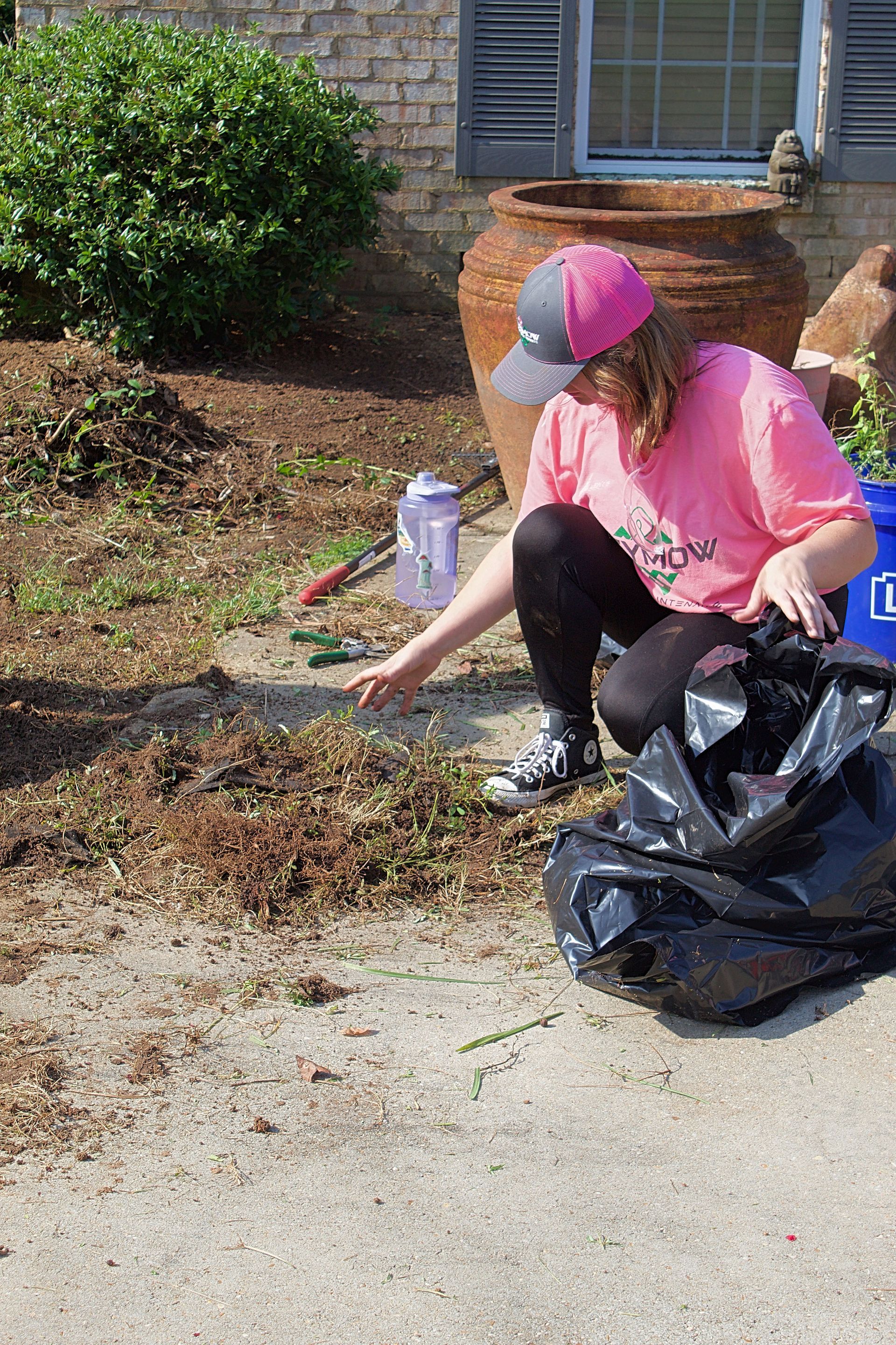 Person in pink shirt and cap crouches by a curb, collecting yard debris into a black trash bag.