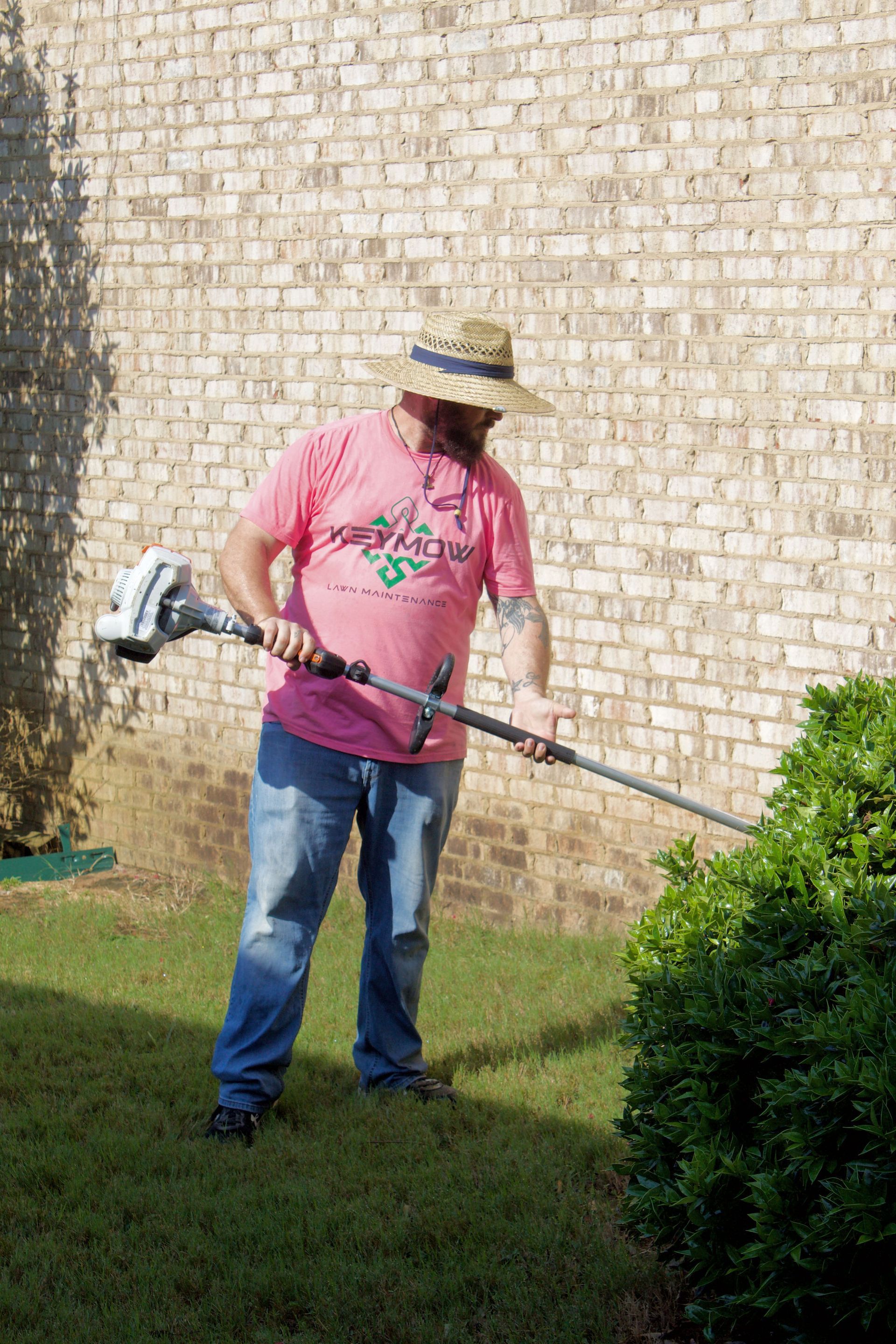 Person using a string trimmer to cut grass near a shrub by a brick wall