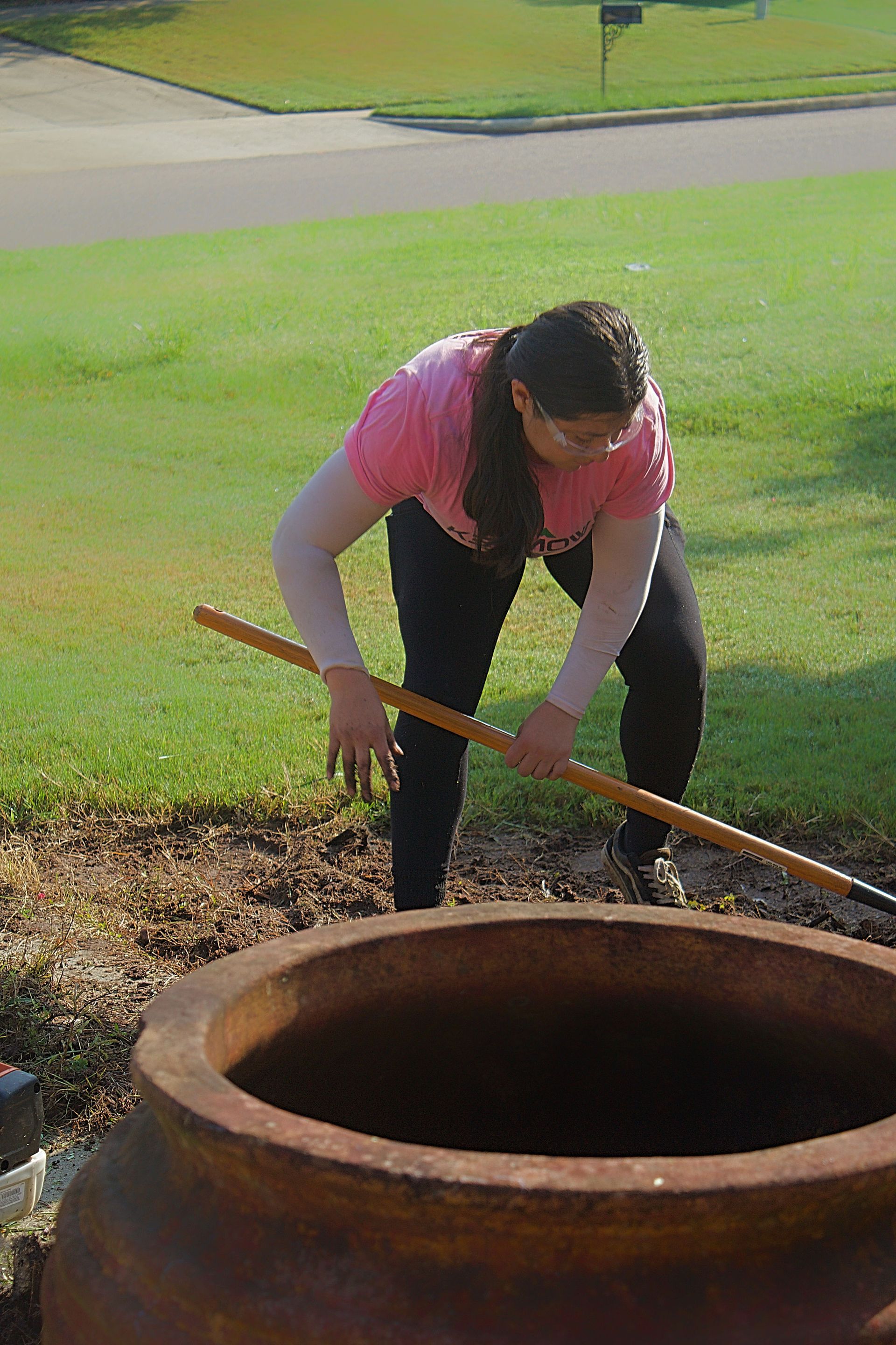 Person in a pink shirt raking soil beside a large clay pot in a grassy yard.