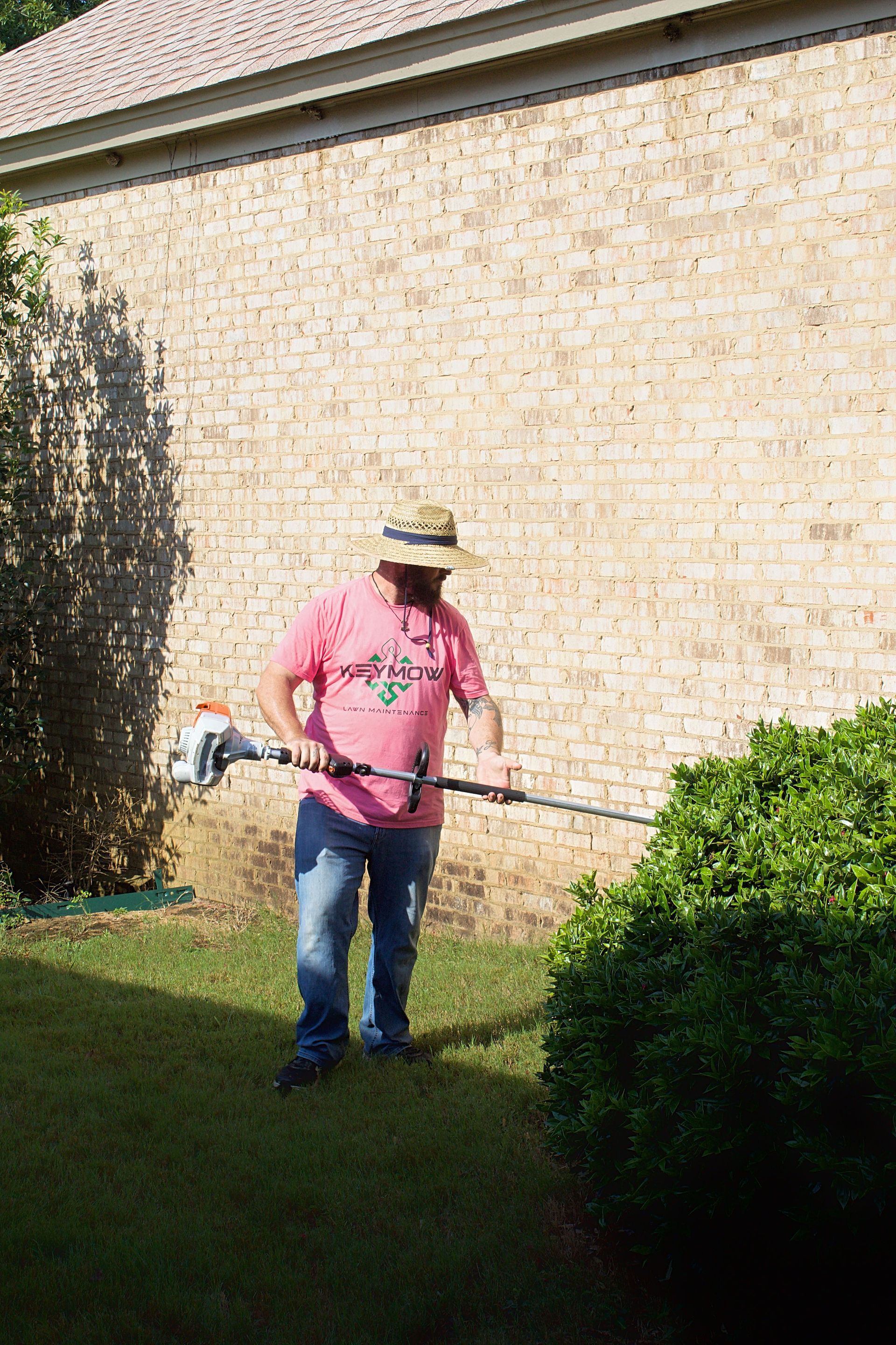 Person using a string trimmer beside a brick wall in a sunny backyard.