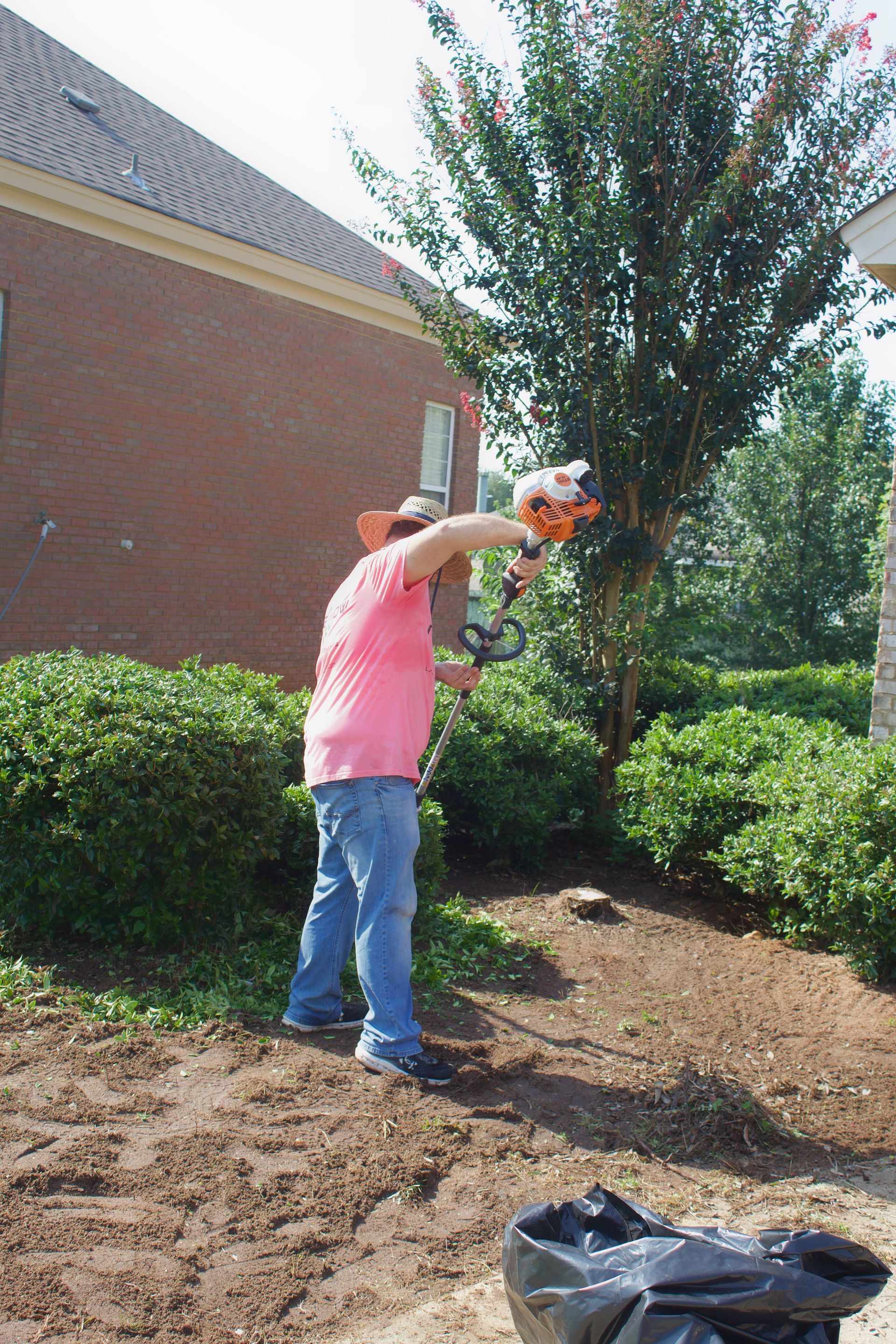 Person using a string trimmer near shrubs beside a brick building, with a tree and dirt foreground.