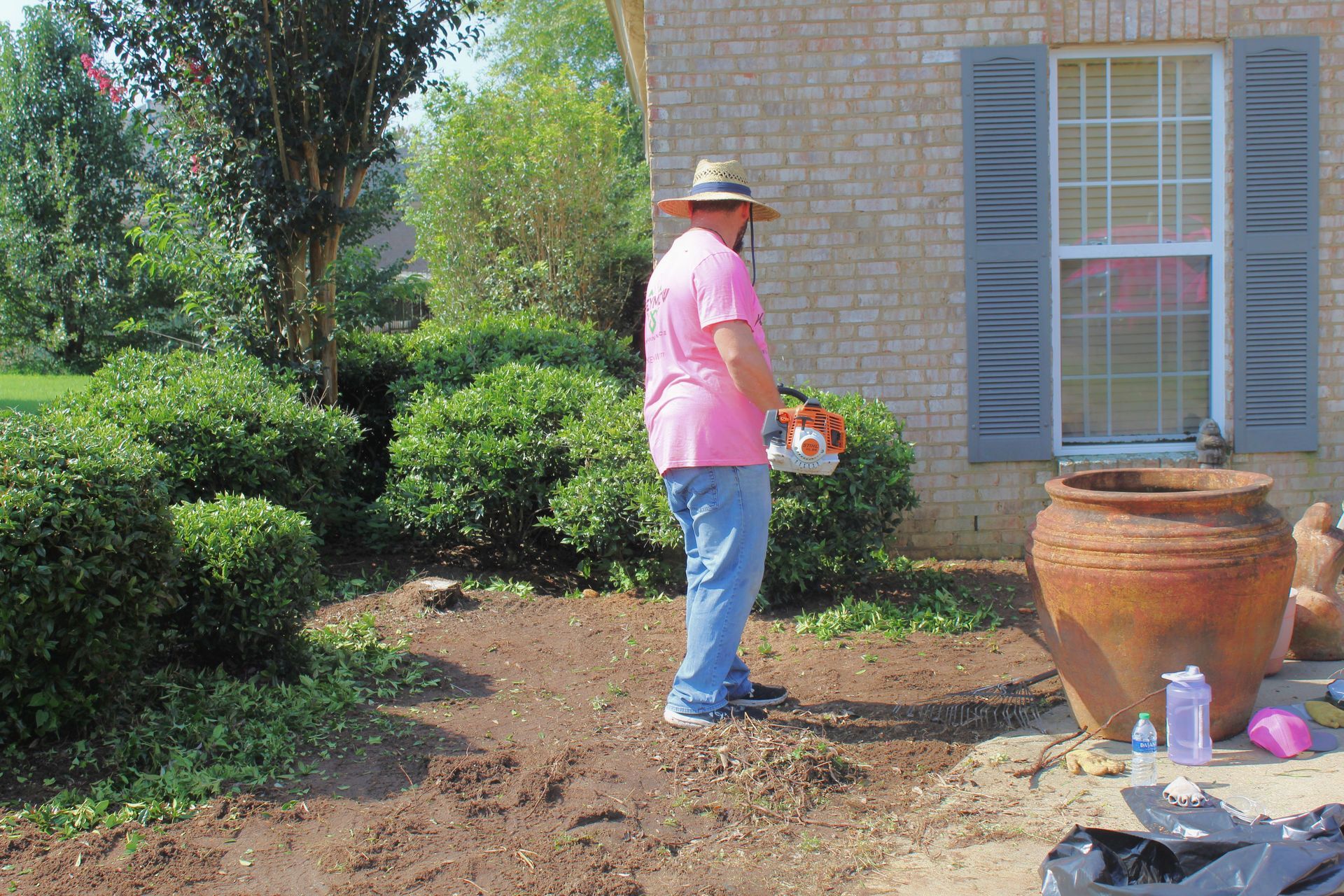 Person in a pink shirt watering plants beside a brick house and large terracotta pot.