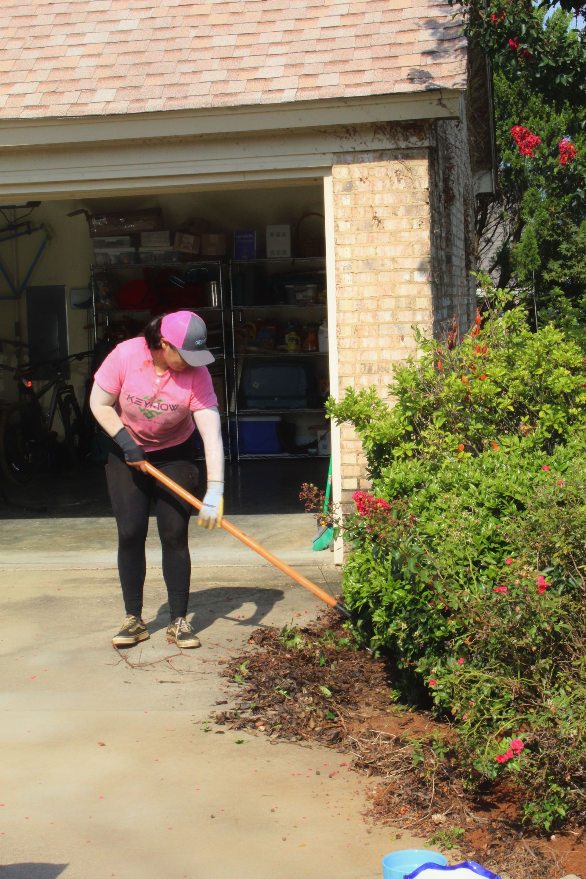 Person raking leaves beside a garage and flowering bushes on a sunny driveway