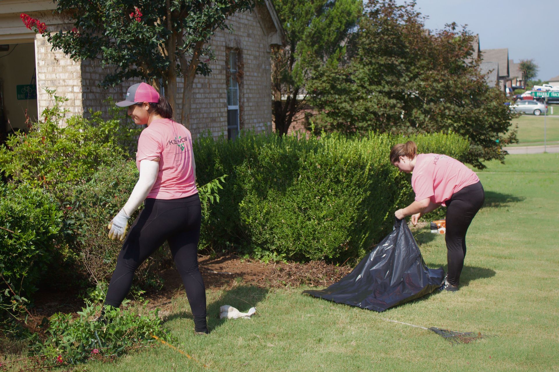 Two people in pink shirts and black pants bagging yard waste on a sunny lawn beside a brick house
