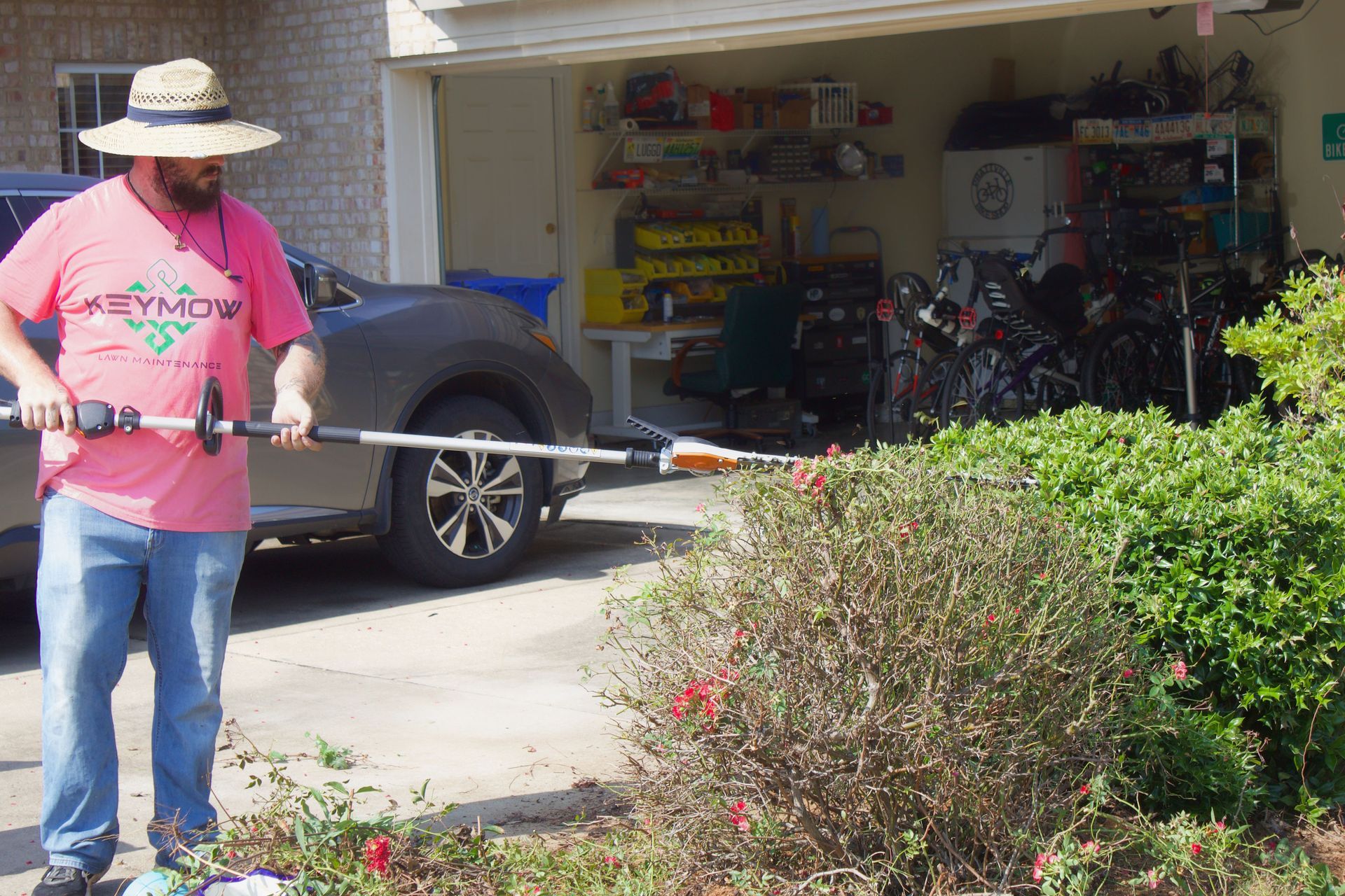 Person in a straw hat using a weed trimmer in a driveway beside a garage and bushes