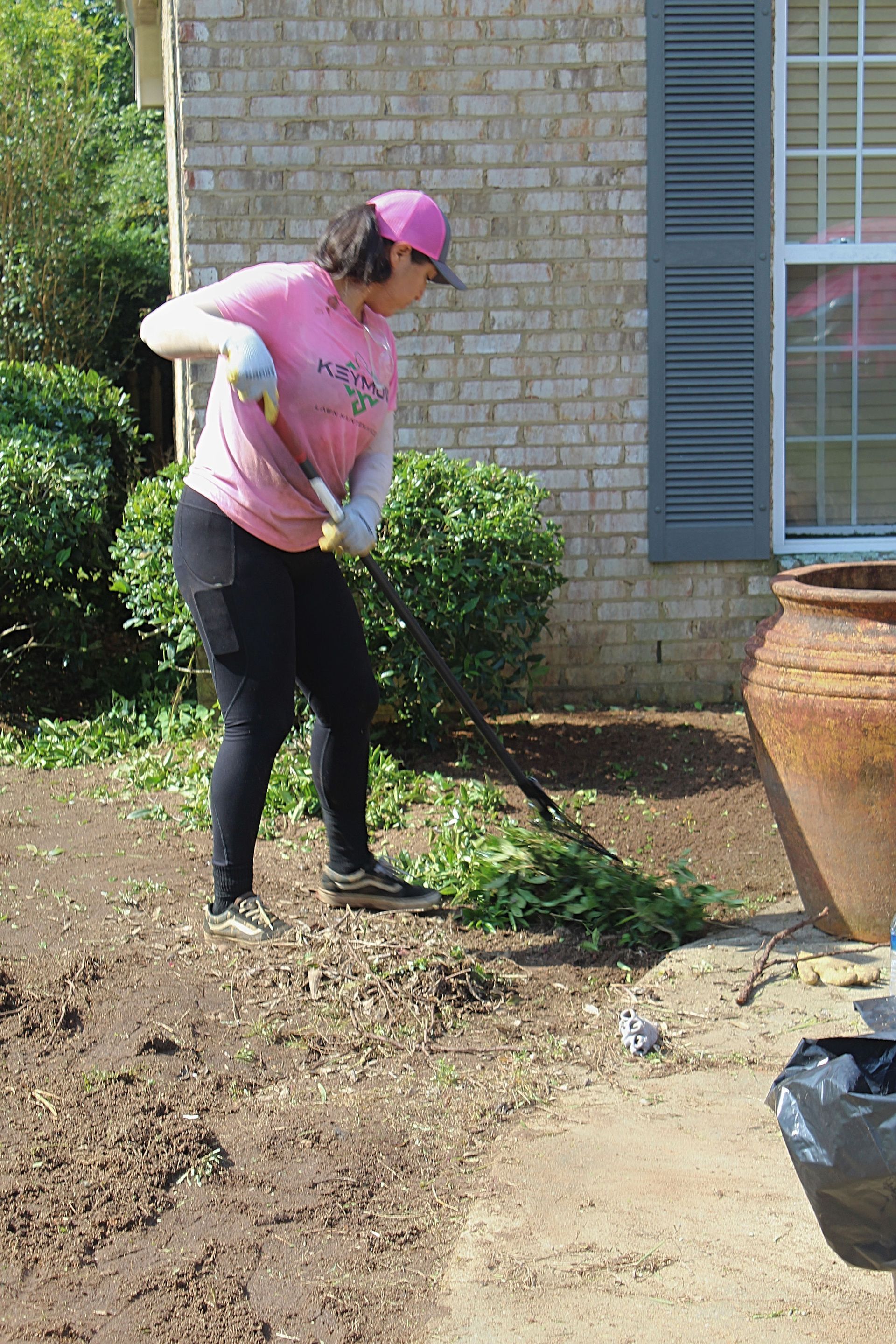 Person in a pink shirt and cap raking soil beside a brick house and window shutters.