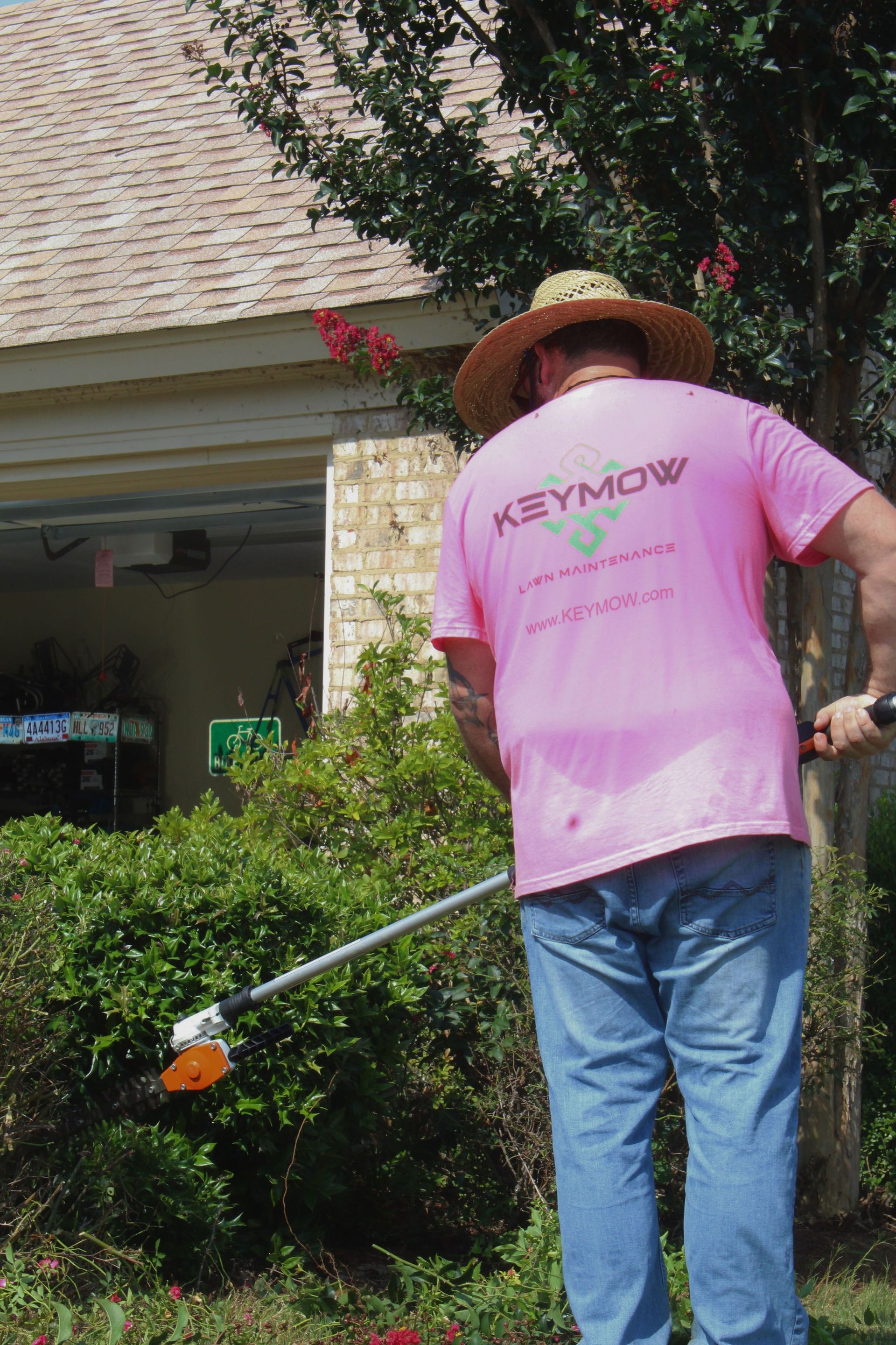 Person in a pink T-shirt and straw hat watering plants in a suburban yard.