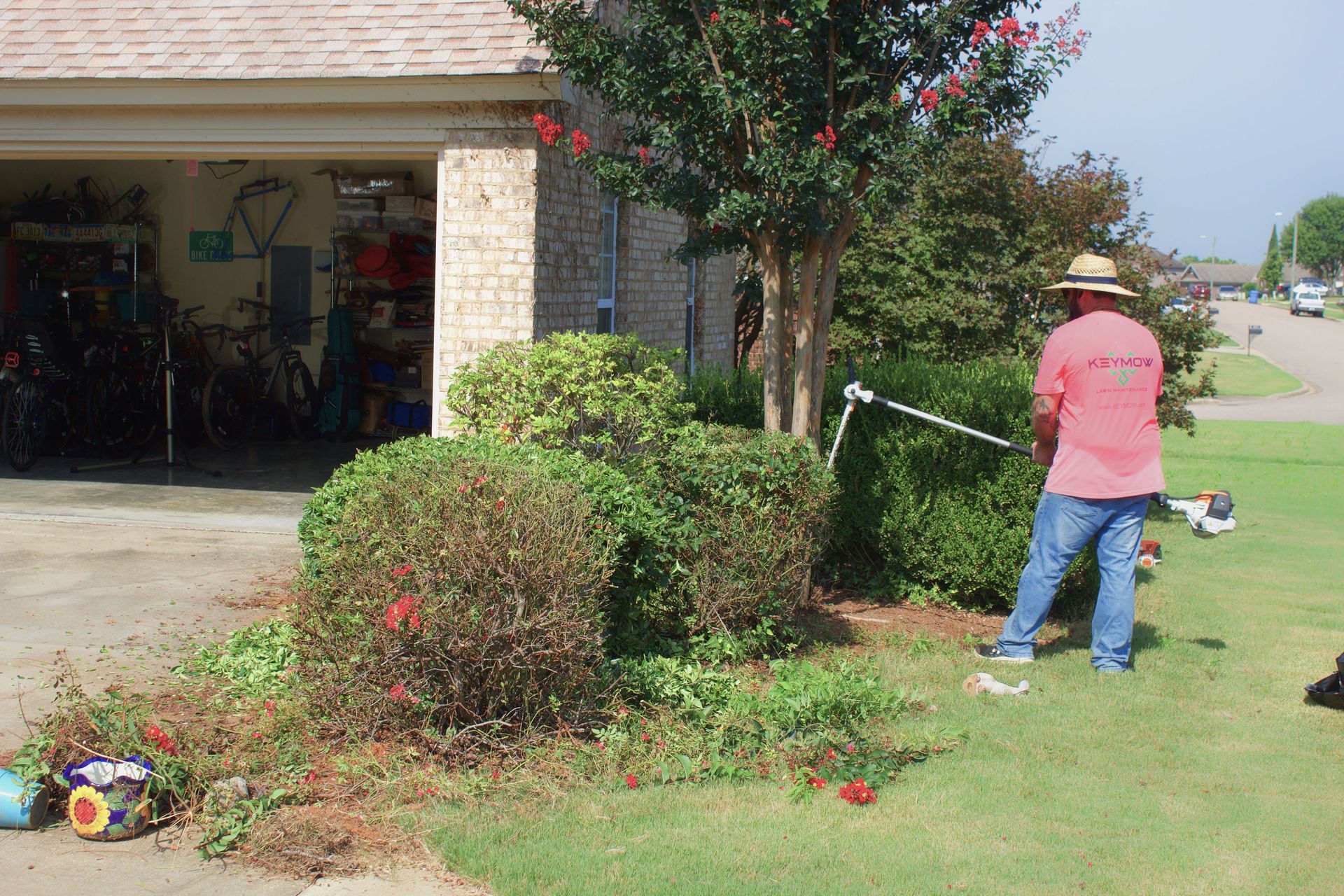 Person in a pink shirt trims bushes with a string trimmer beside a suburban garage.