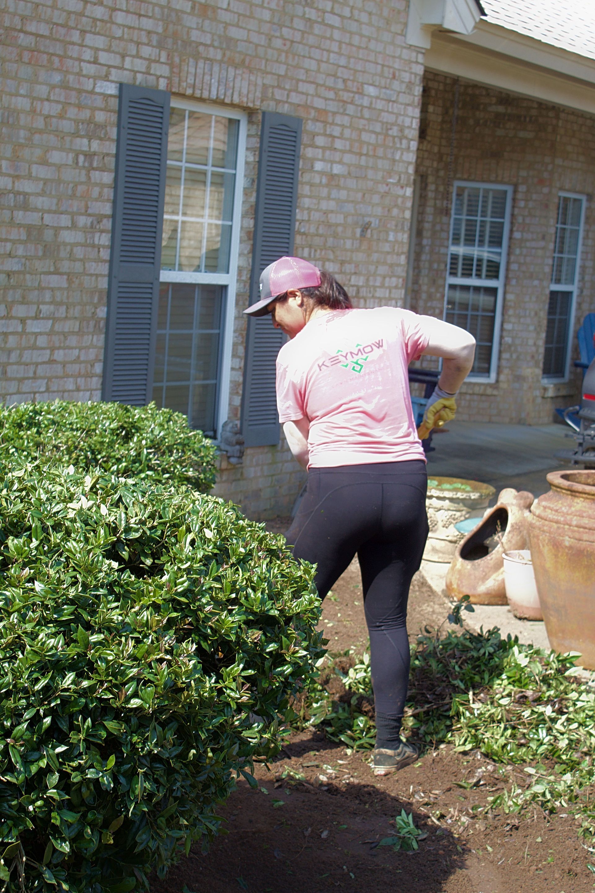 Person in pink cap and shirt gardening beside a brick house, trimming bushes.
