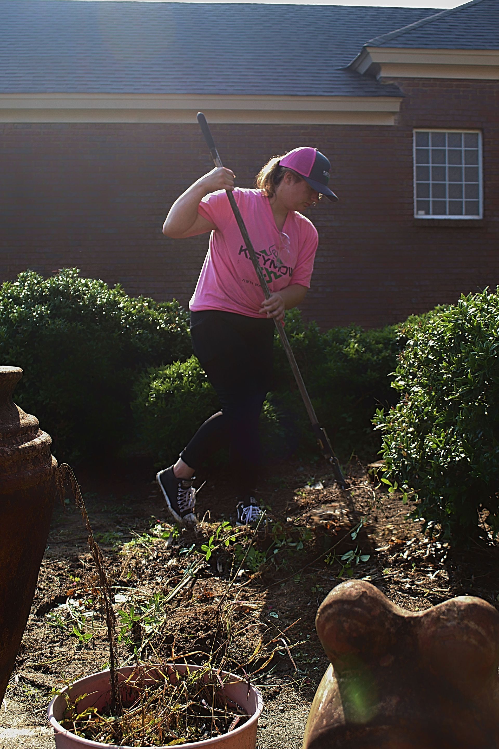Person in a pink shirt and cap gardening with a shovel in a yard near a brick house.