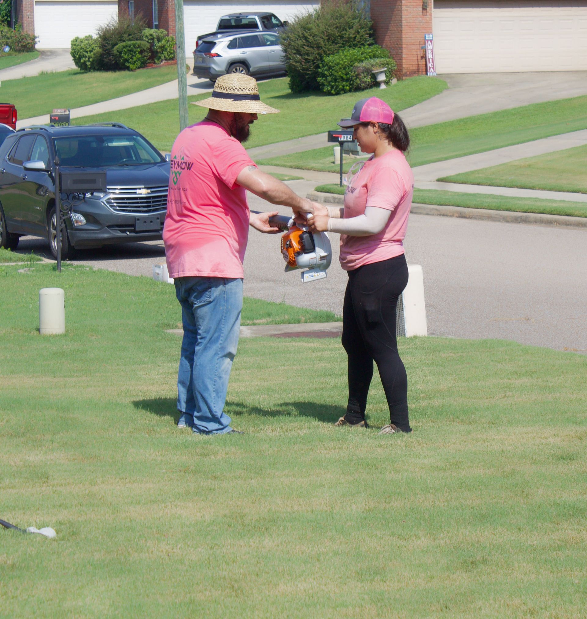 Two people in pink shirts exchanging a tool on a suburban lawn near parked cars.