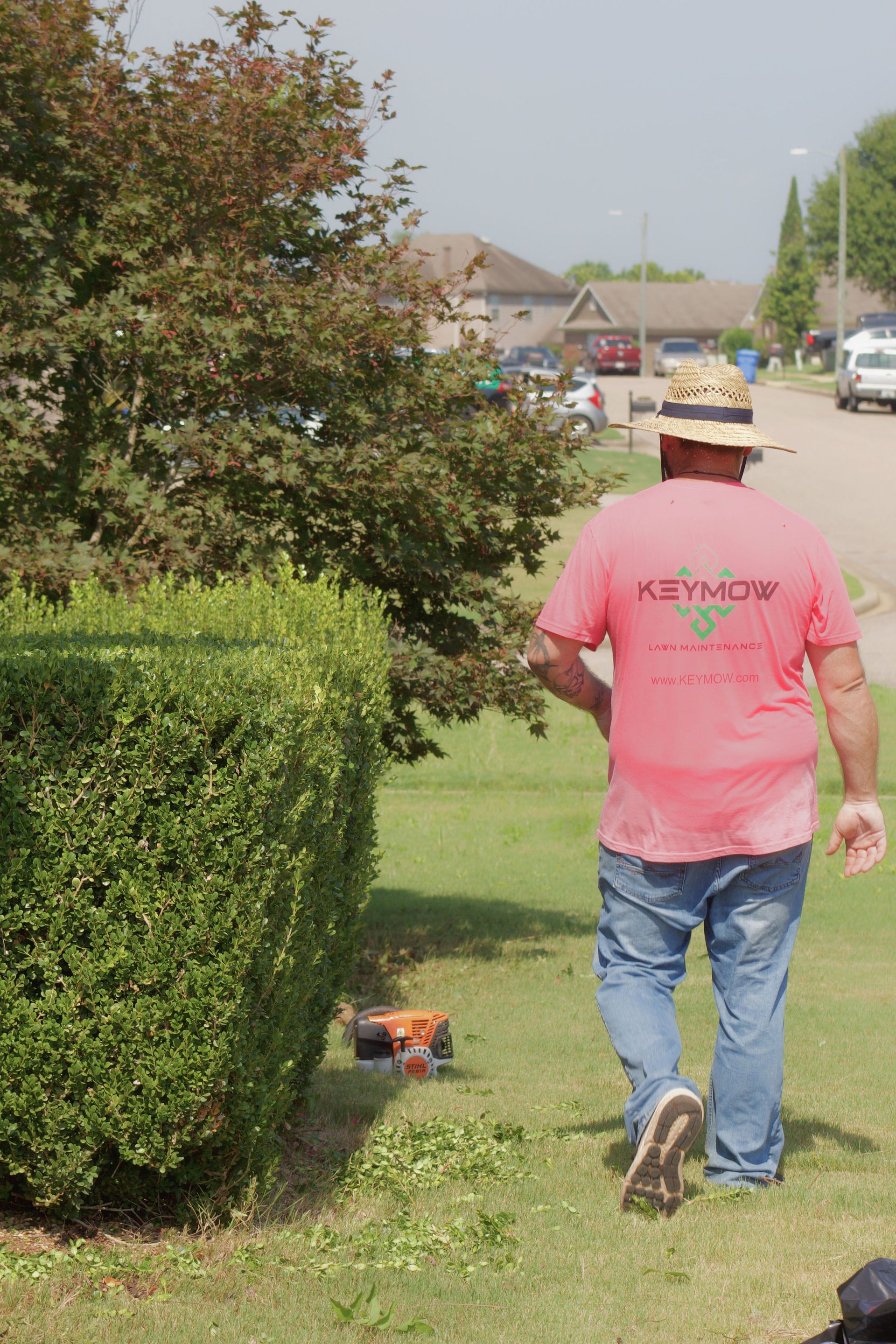 Person mowing a suburban lawn beside hedges, wearing a pink shirt and straw hat.