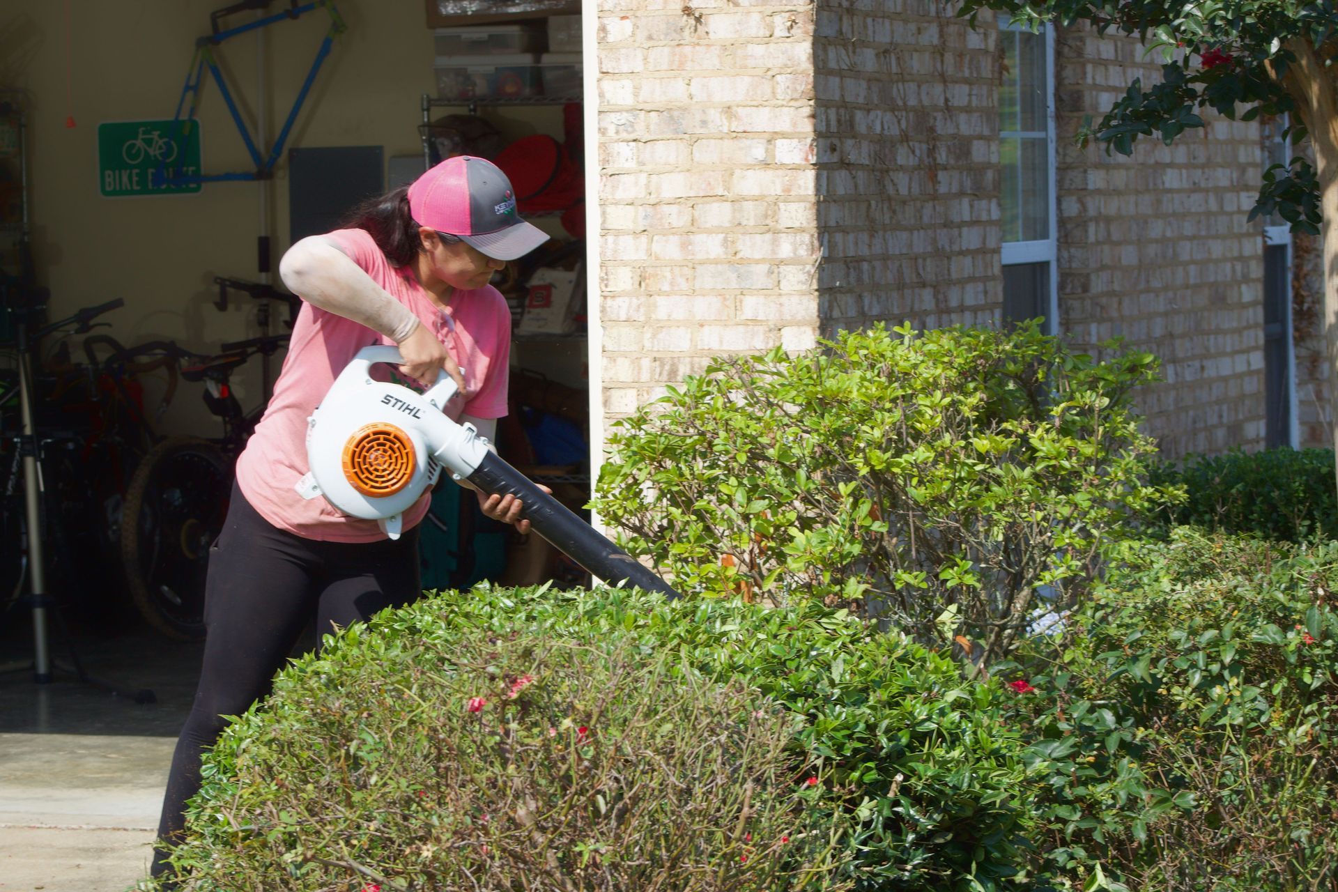 Person using a leaf blower to clear bushes beside a brick building