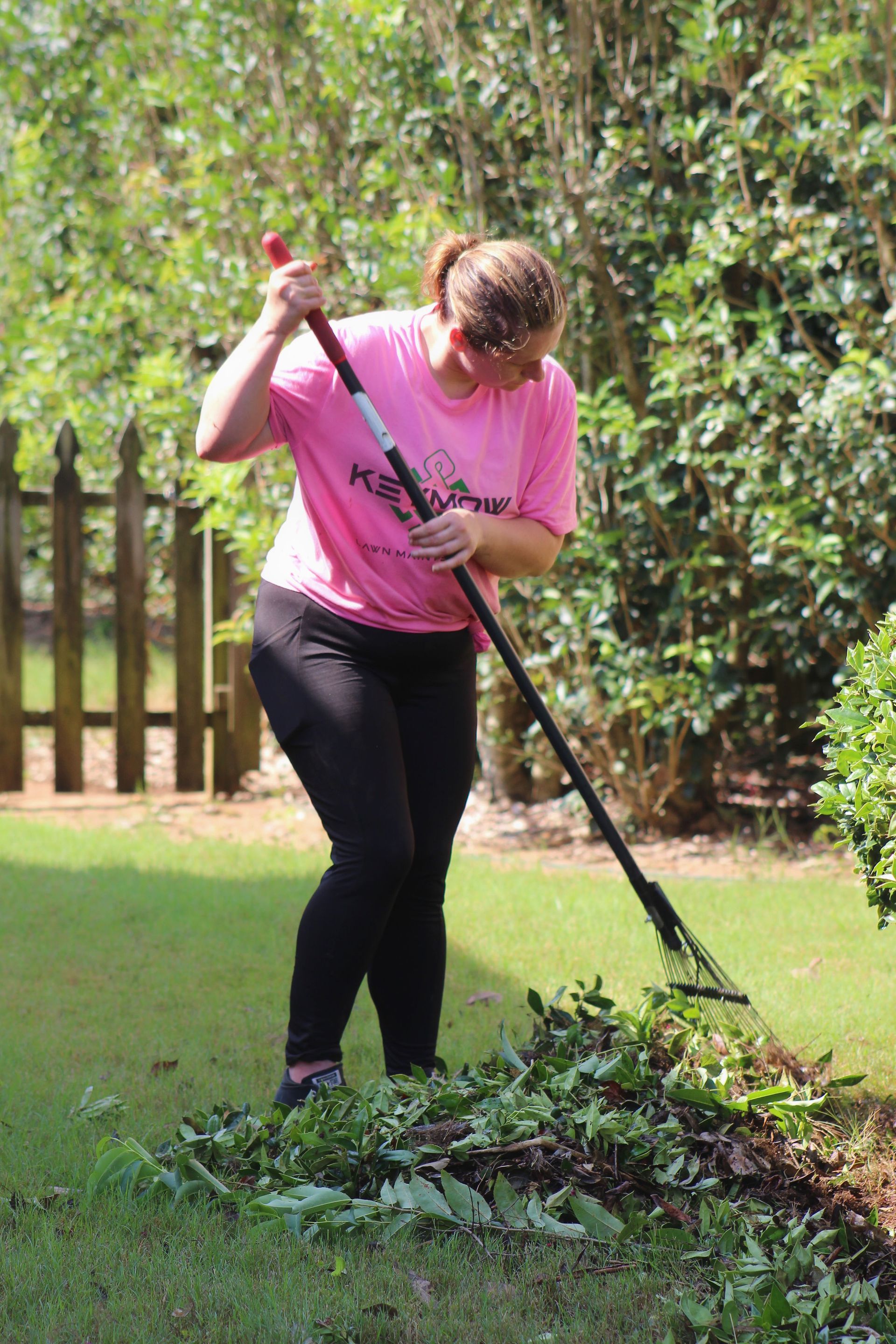 Person in a pink shirt rakes leaves in a grassy yard beside a wooden fence.