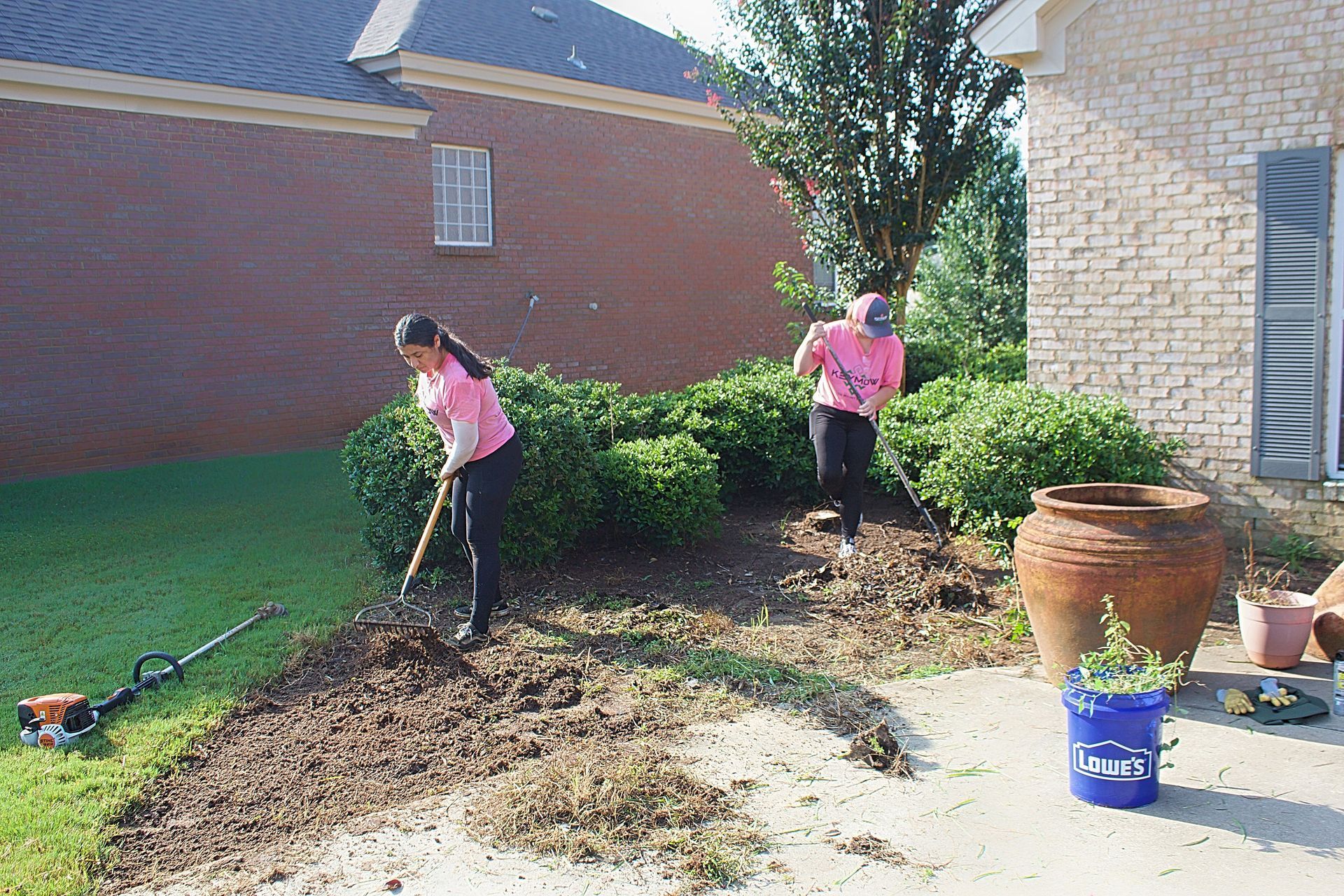 Two people in pink shirts raking leaves in a yard beside a brick house