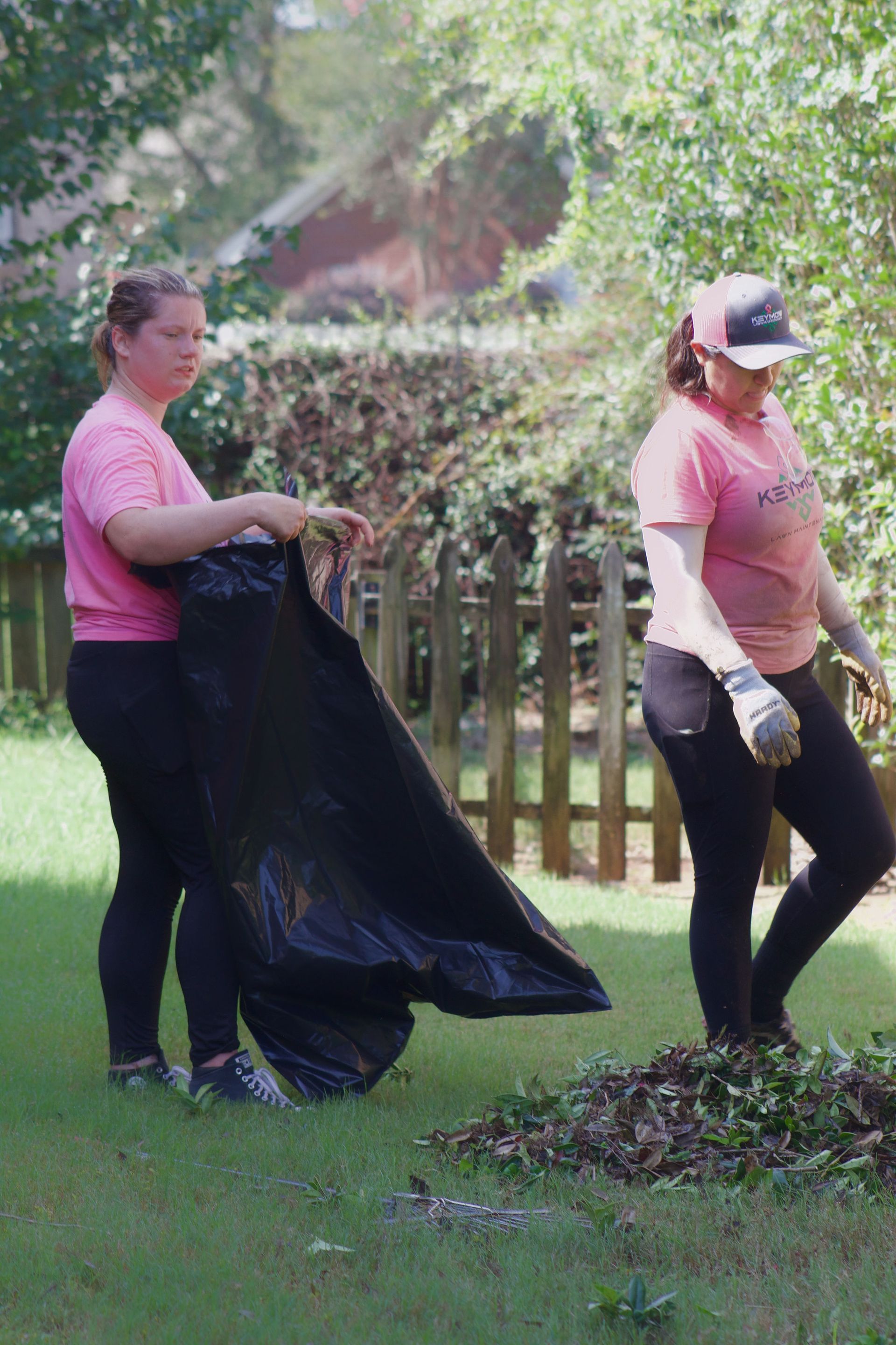 Two people in pink shirts cleaning up leaves in a grassy yard with a black bag and rake