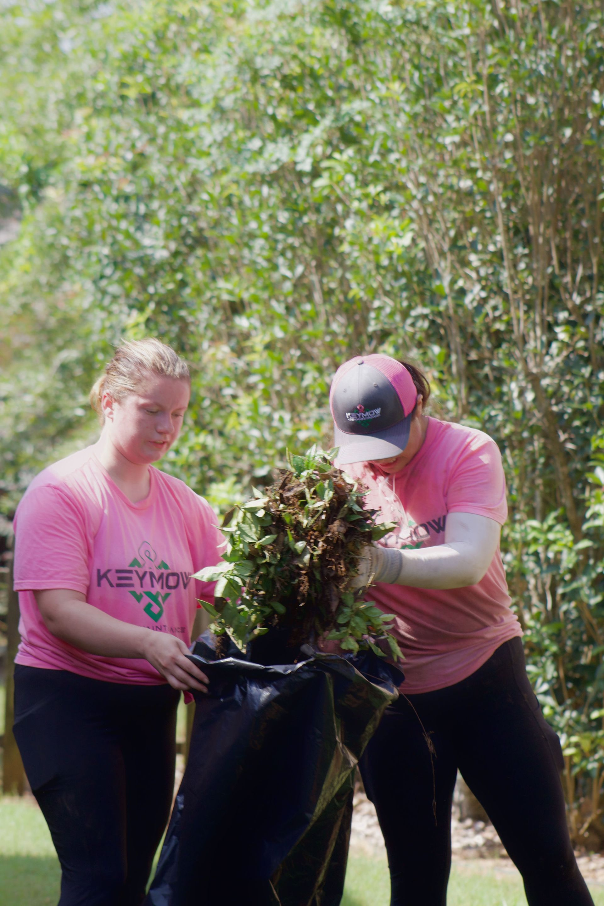 Two people in pink shirts gardening together, lifting a plant from a black bag outdoors.