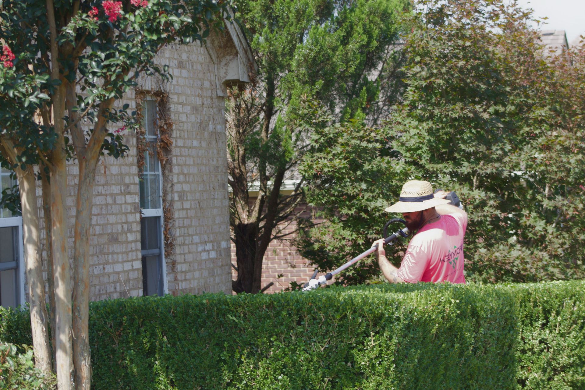 Person in a straw hat using a string trimmer beside a hedge near a stone house