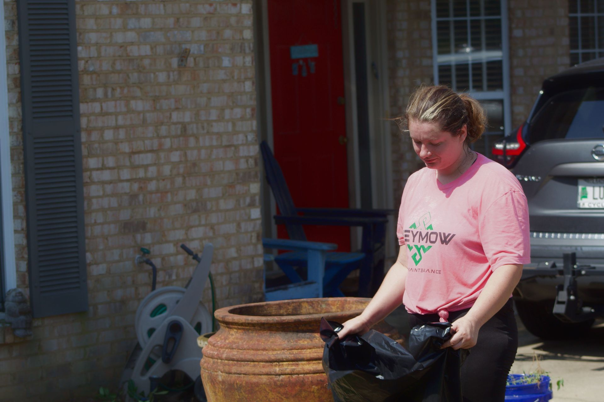 Person in a pink shirt standing beside a large planter outside a brick house