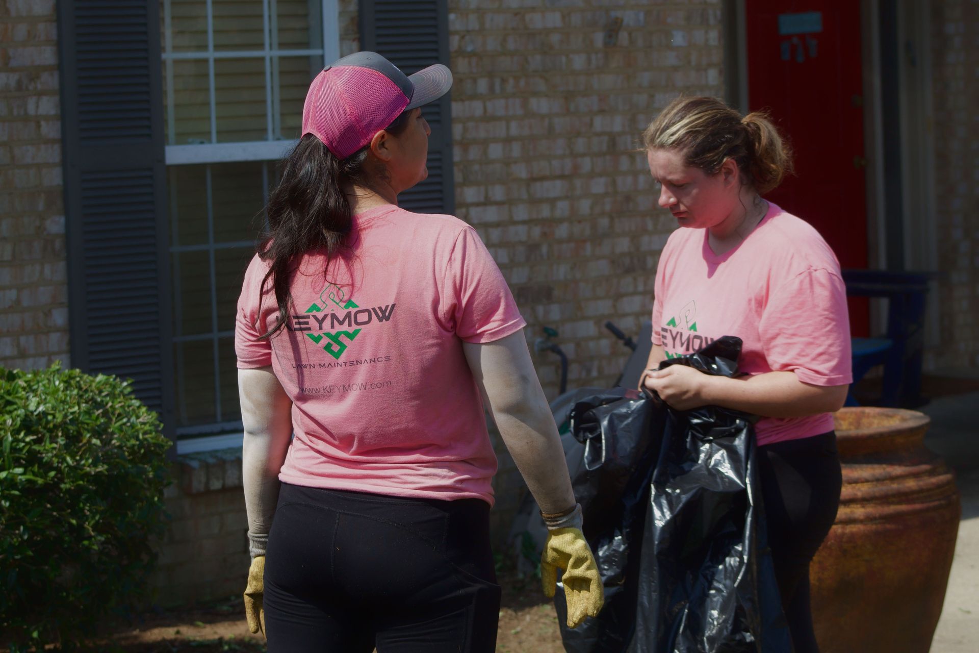 Two volunteers in pink shirts and gloves sorting items outdoors beside a brick building.