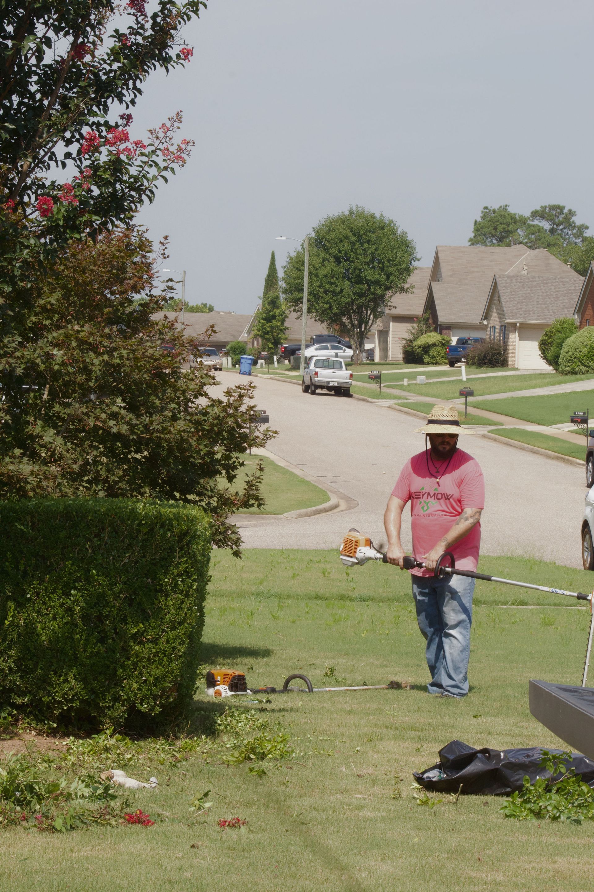 Person using a string trimmer on a suburban front lawn with a hedge and houses in the background