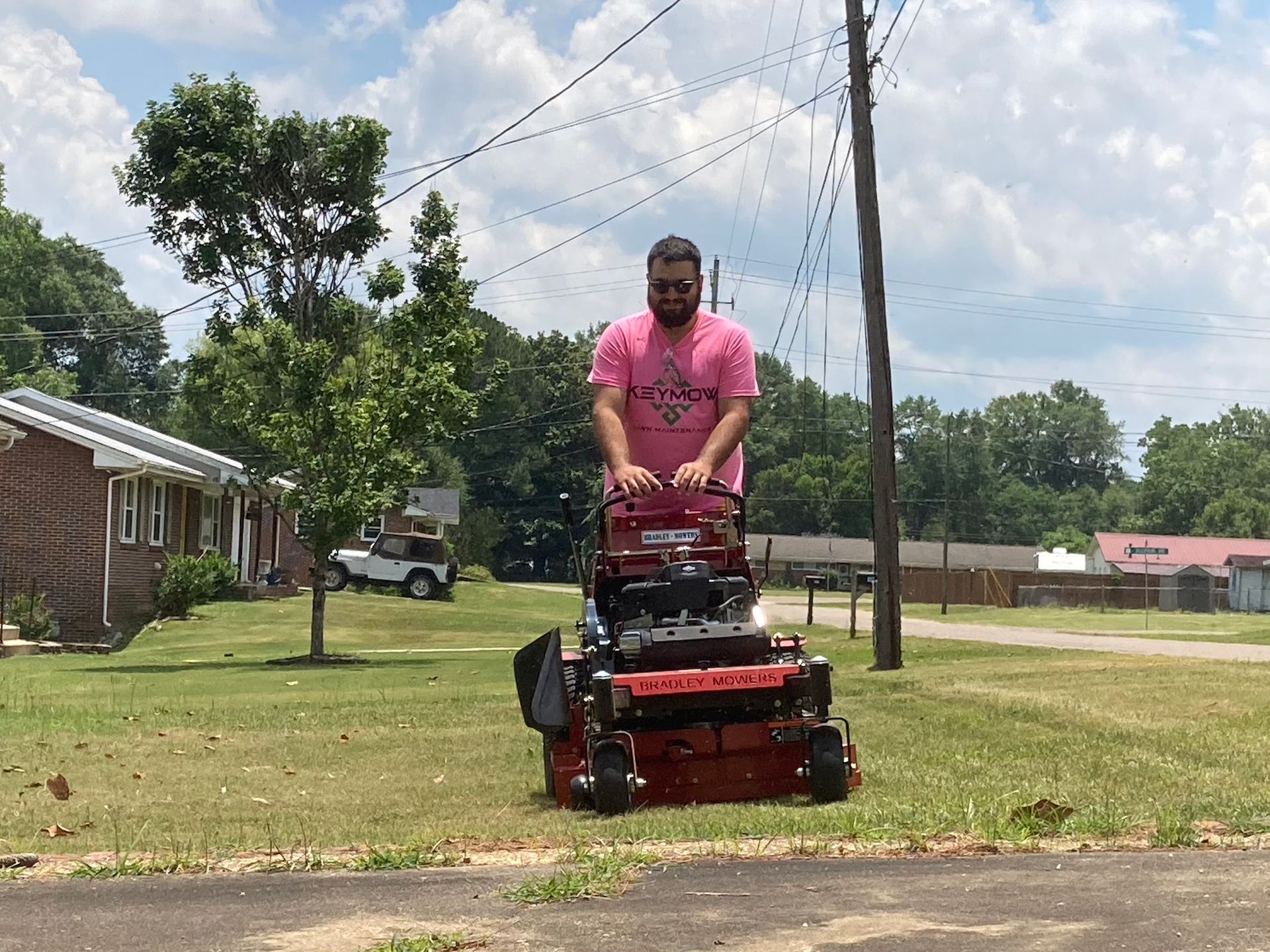 Person in a pink shirt riding a red lawn mower on a grassy yard near houses and trees.