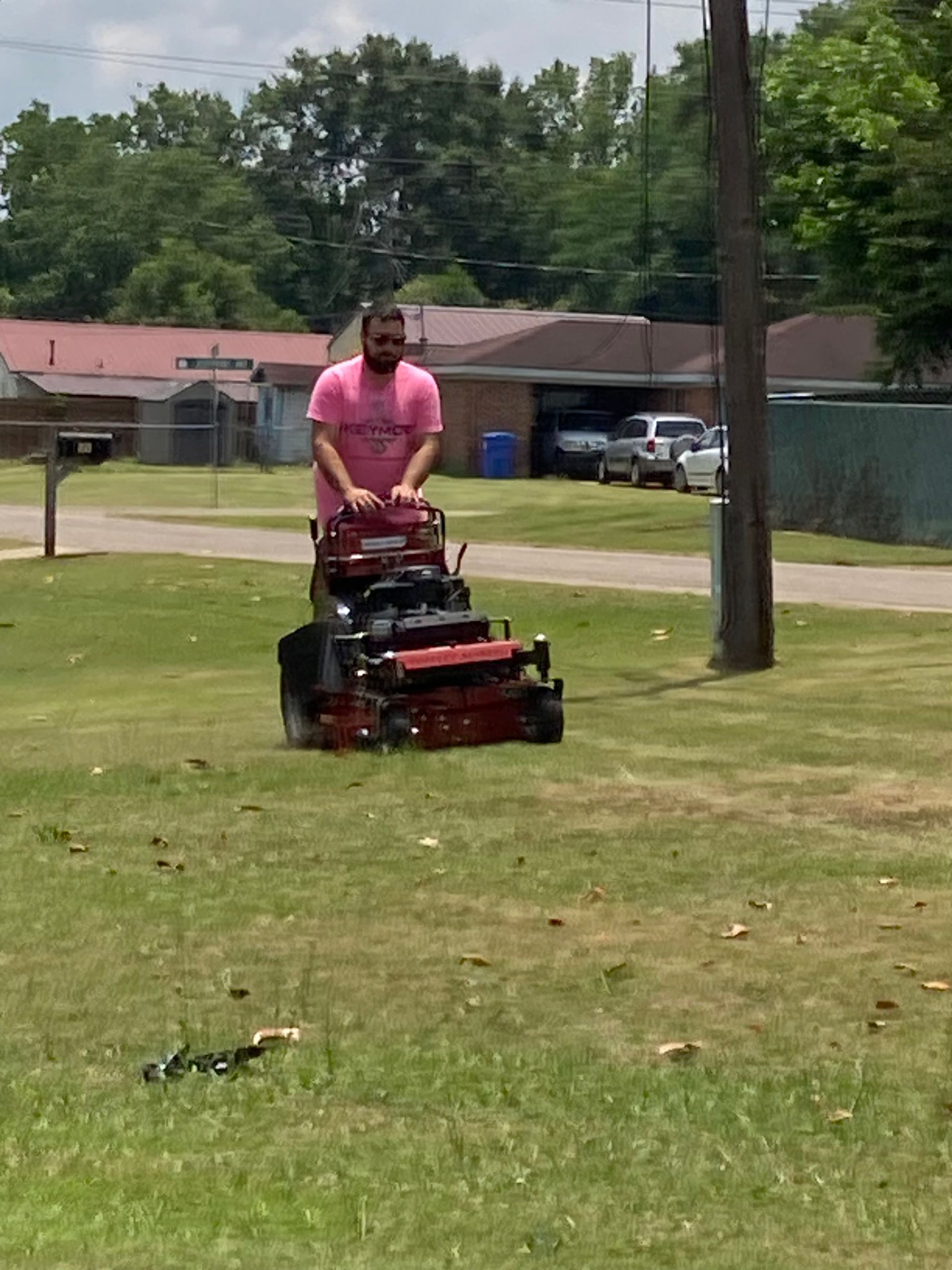 Person in a pink shirt mowing a grassy lawn with a red mower in a residential area.