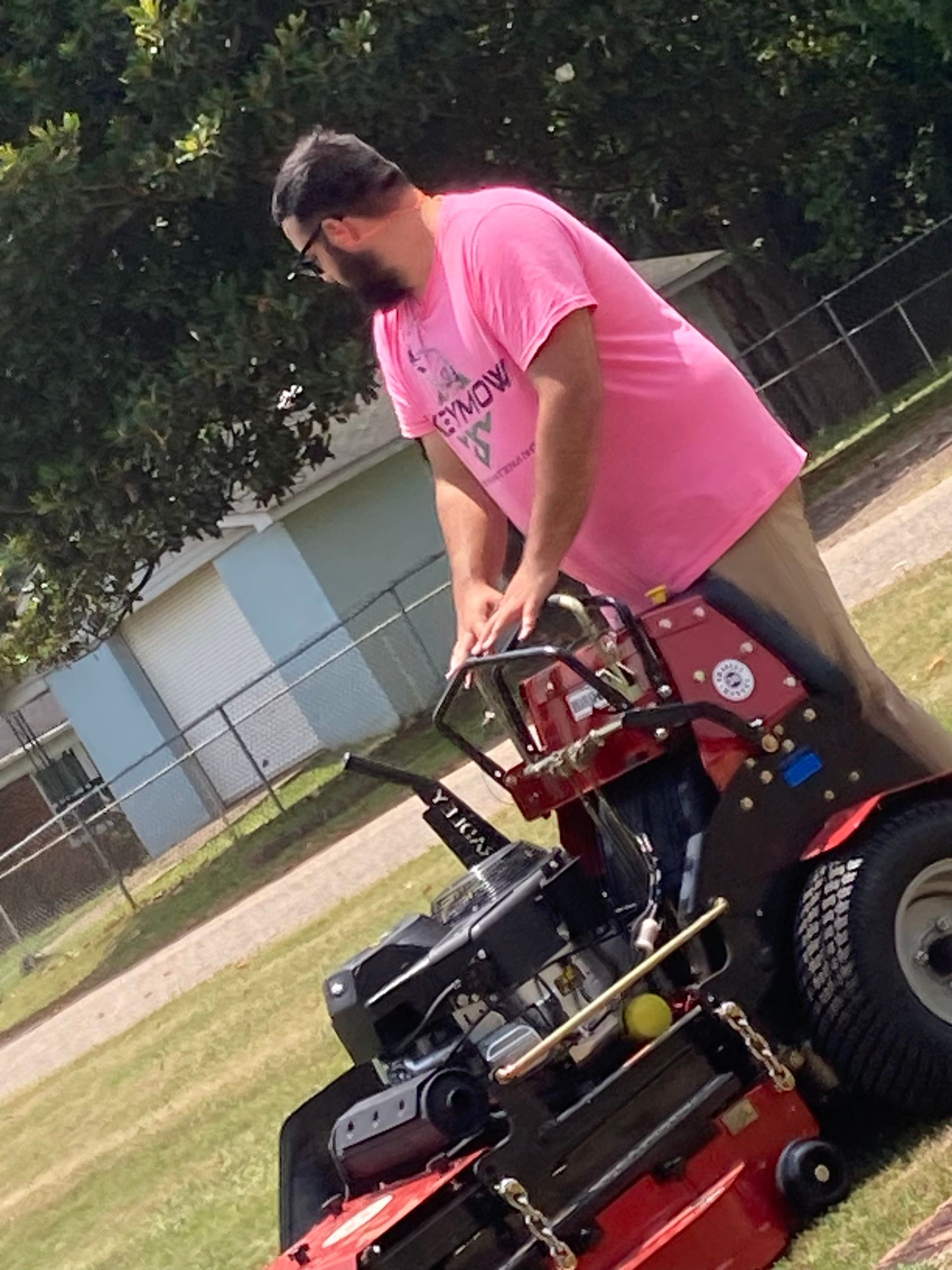 Man in a pink shirt riding a red zero-turn lawn mower on a grassy field
